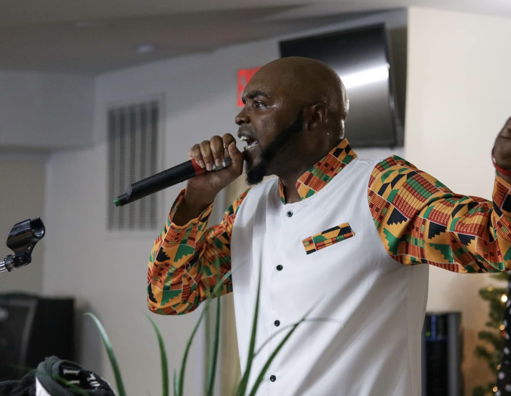 Man singing into a microphone, wearing a colorful patterned shirt and a white vest, indoors.