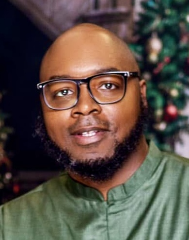 Man with glasses and beard, wearing a green shirt, standing in front of a decorated Christmas tree.