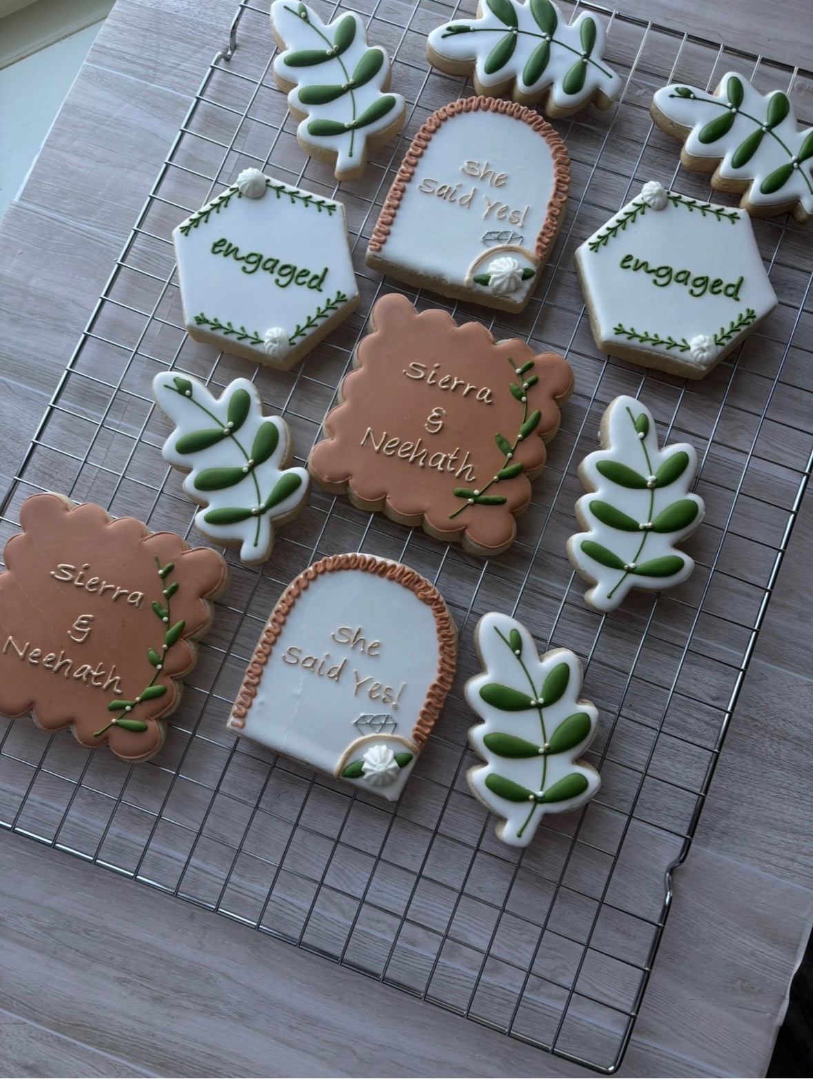 Decorative cookies on a wire rack, including leaf-shaped, hexagon-shaped with 'engaged' text, and arch-shaped with names, in neutral colors.