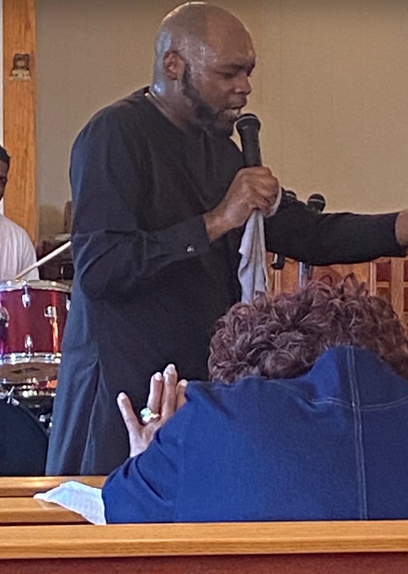 Man holding microphone speaking to audience in indoor setting, with drum set visible in background.