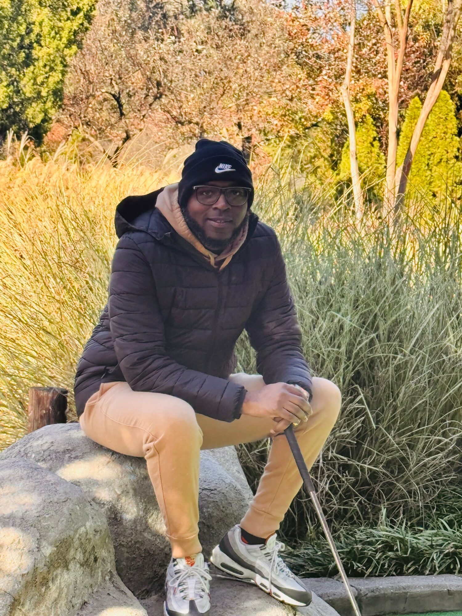Person sitting on a rock outdoors surrounded by tall grass and trees, wearing a black jacket, beige pants, and a black beanie.