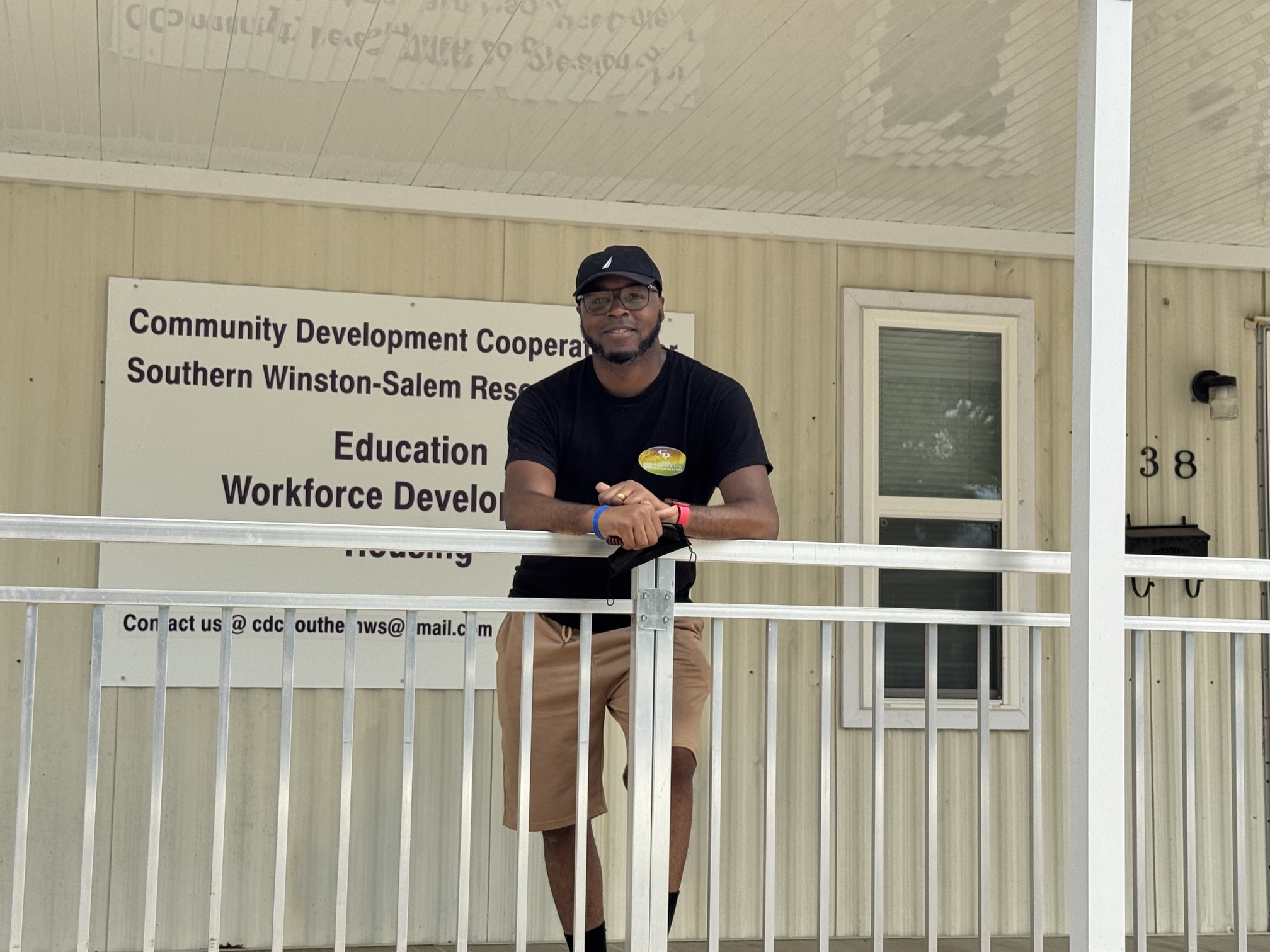 Man standing outside a building with a sign, wearing a black shirt, beige shorts, and a cap.