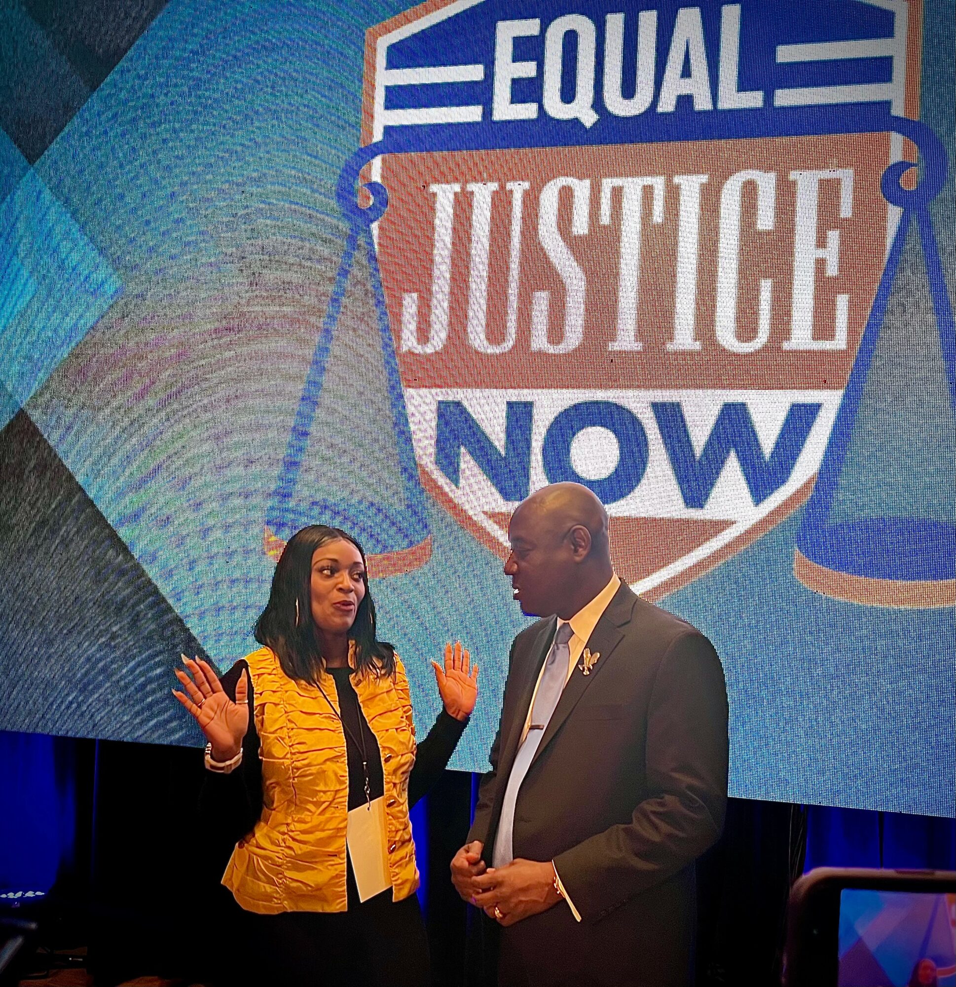 Two people speaking on stage with a large screen behind them displaying a sign that reads 'EQUAL JUSTICE NOW'.