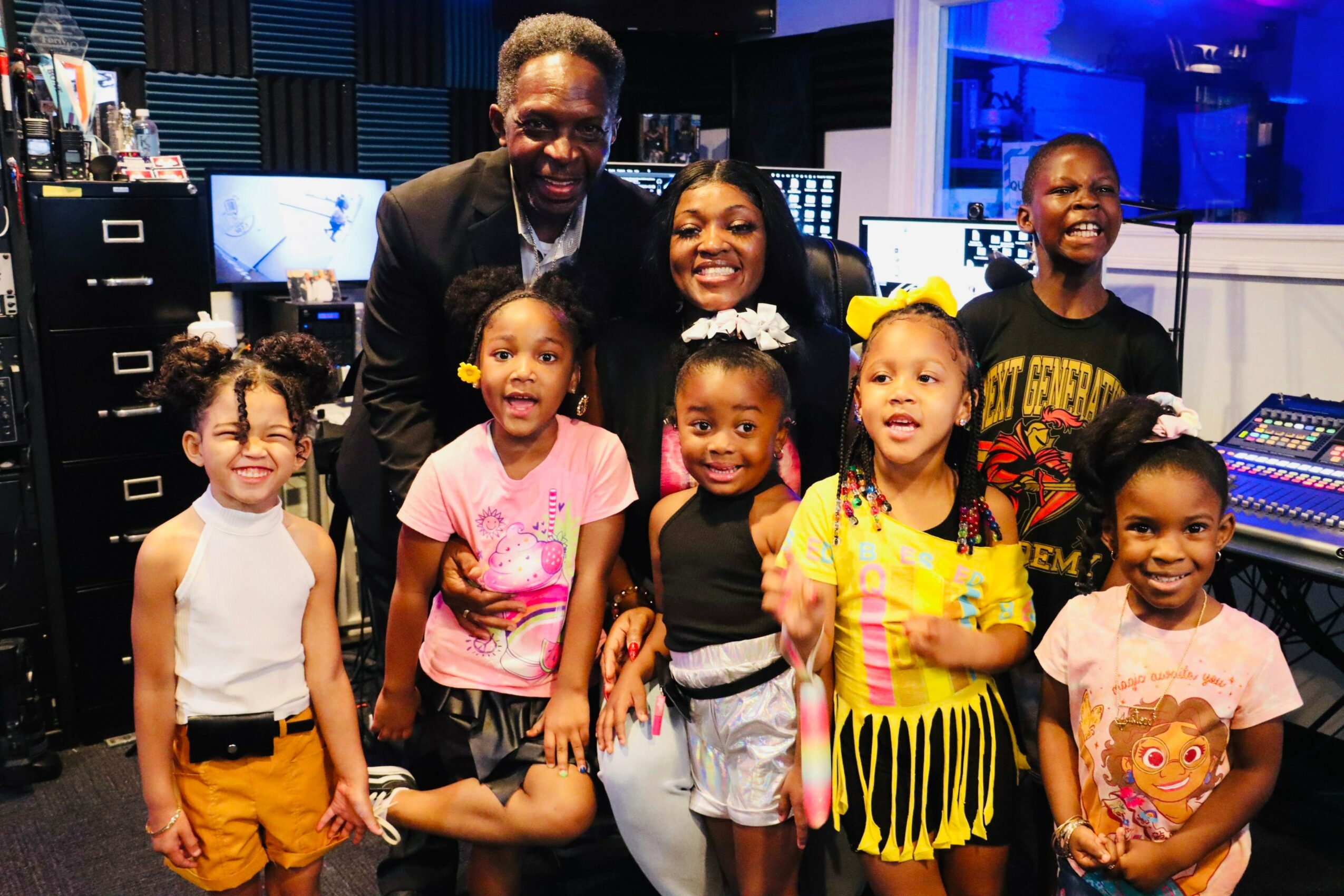 Group of children and two adults smiling in a recording studio with computers and sound equipment.