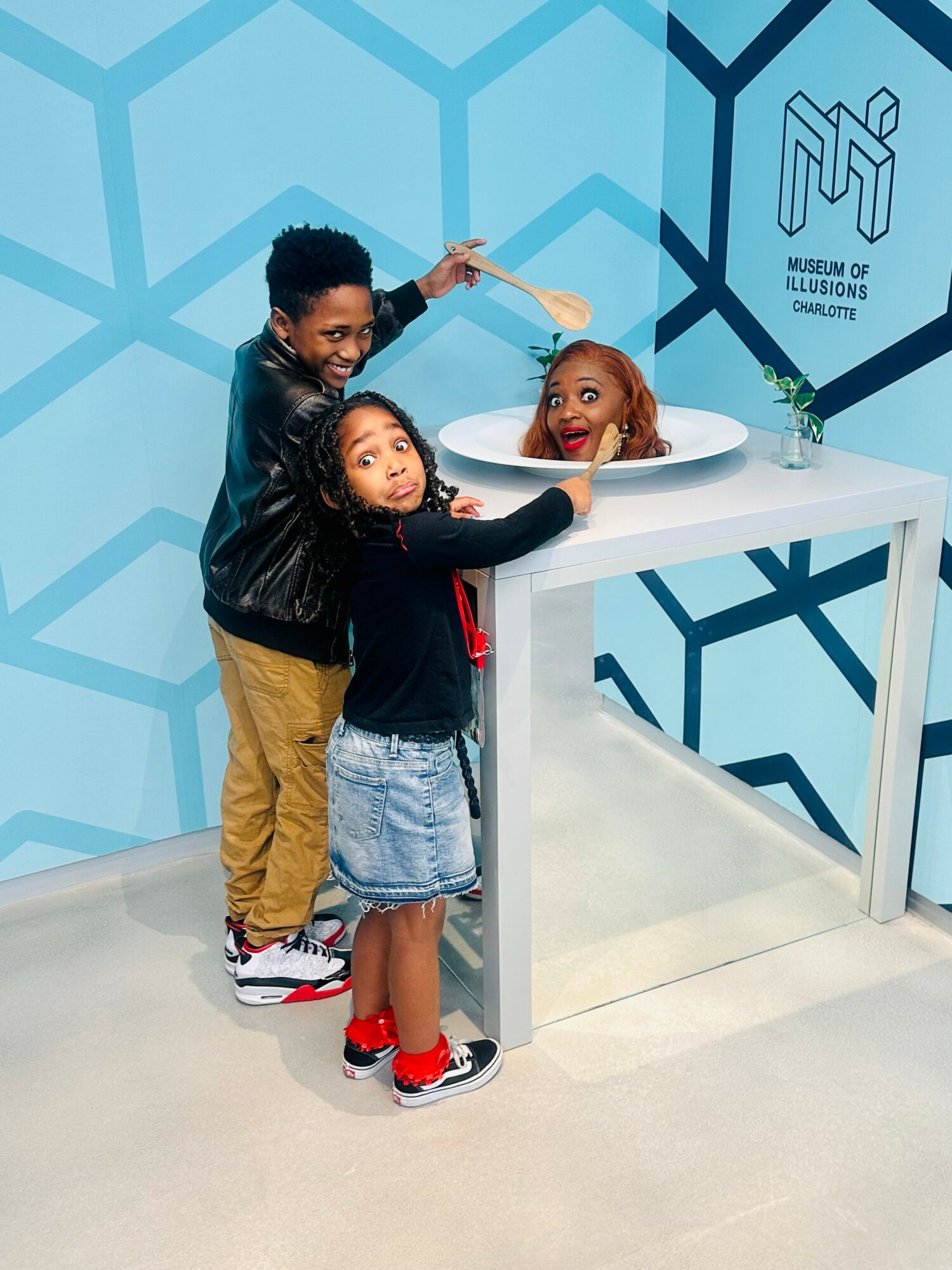 Three children at an interactive exhibit, with one child in a bowl and two children touching the bowl, in a room with blue geometric wall pattern.
