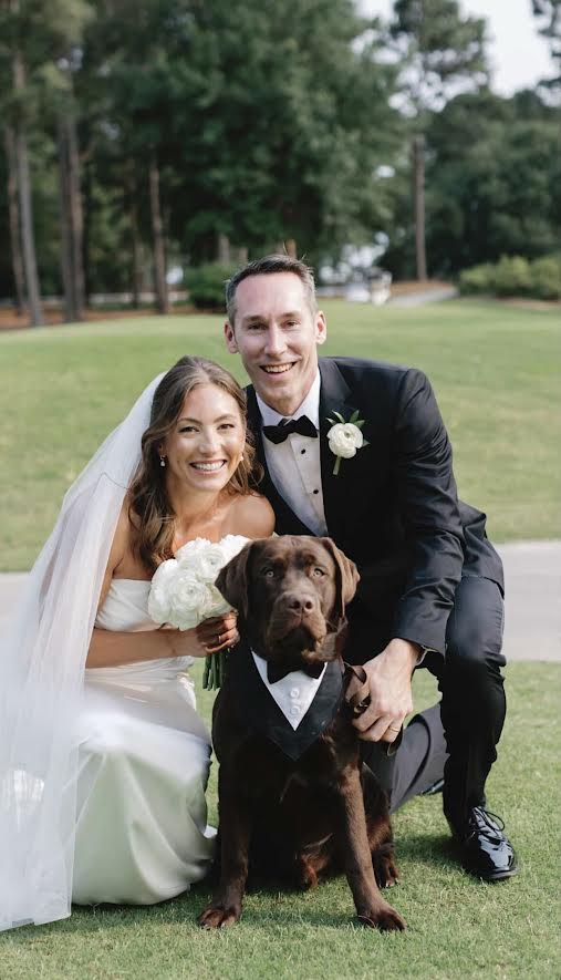 Bride and groom with a brown dog outdoors on grass, trees in background, smiling, wedding attire, bride holding bouquet.