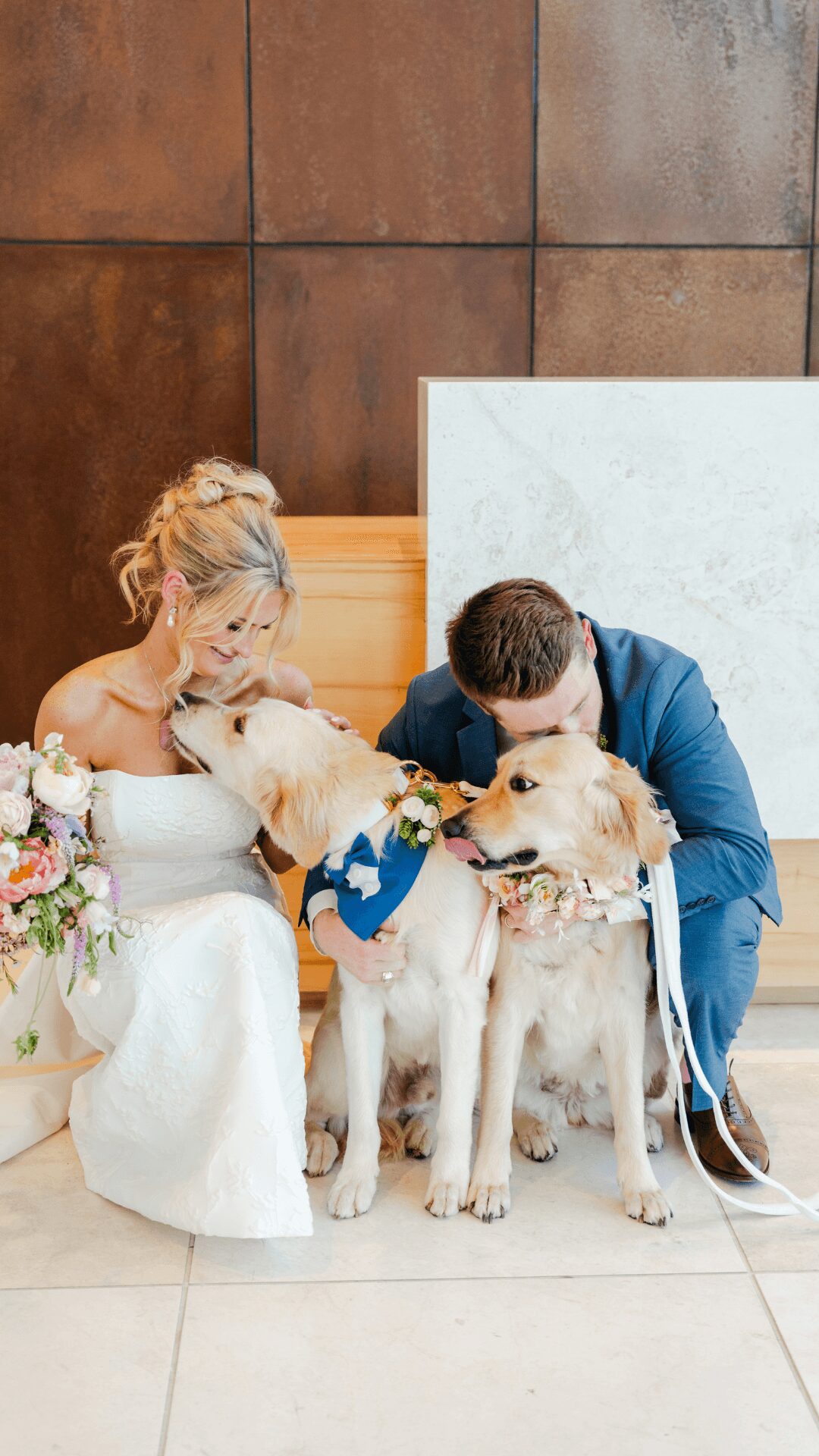 Bride and groom with two dogs, one wearing a blue bandana, in front of a tiled wall and white marble surface.