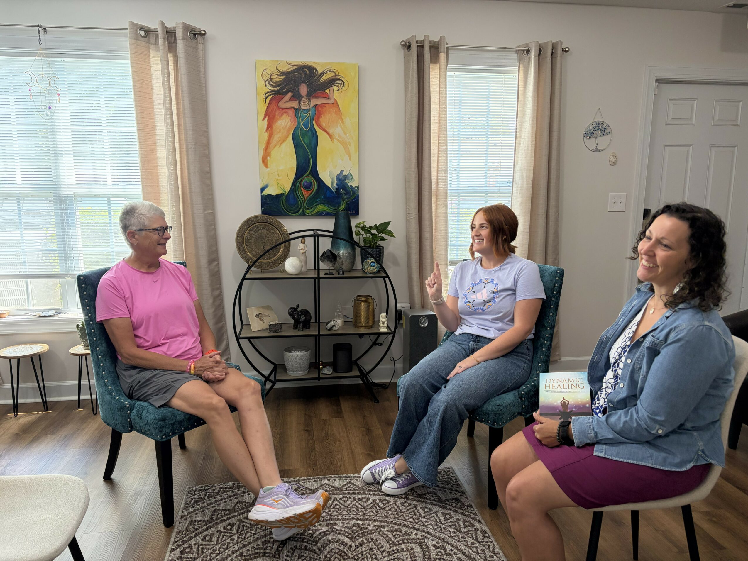 Three women sitting and talking in a living room with large windows, curtains, a painting, and a shelf with decor.