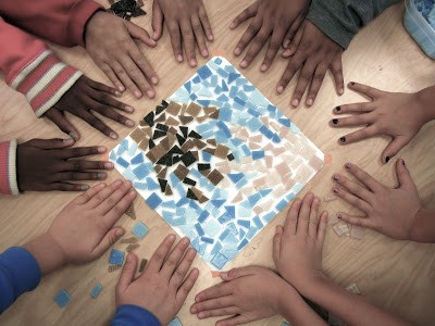 Multiple hands surround a mosaic artwork on a table, with various skin tones and clothing styles visible.