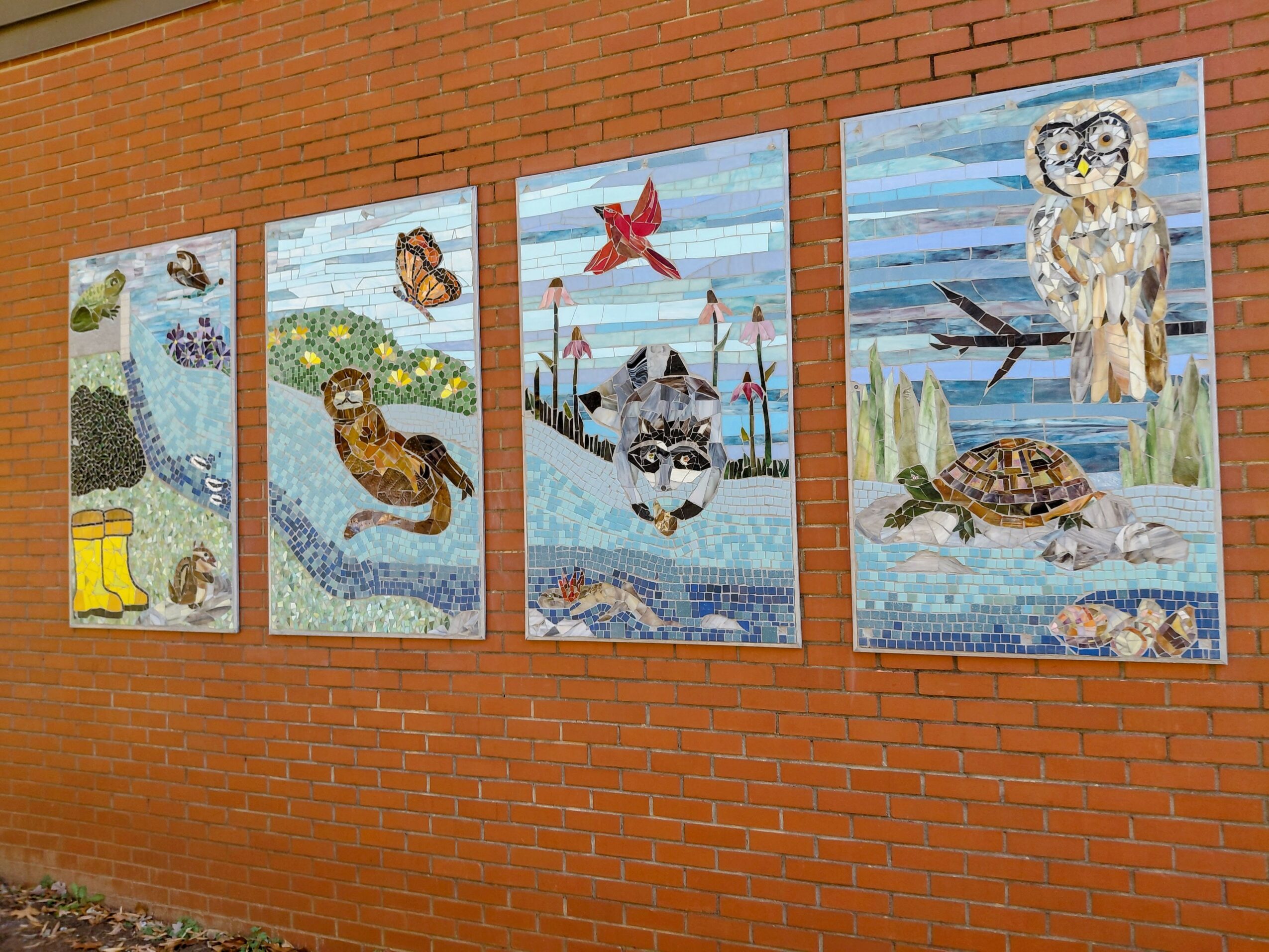 Four colorful murals of animals and nature scenes on a brick wall, including a fox, owl, turtle, and a bird in a pond.