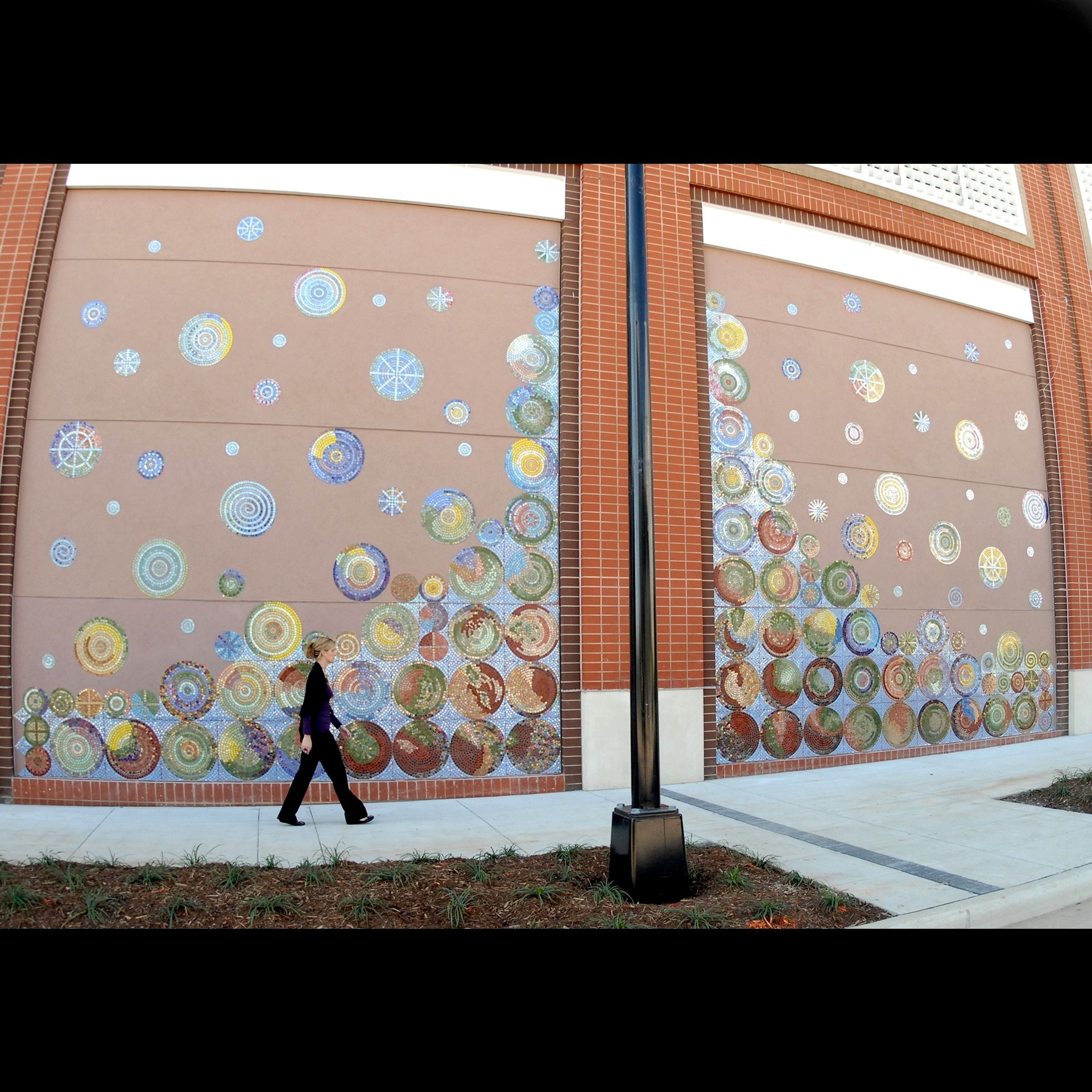 Person walking past a large window decorated with colorful circular patterns, brick building, sidewalk, and grass.