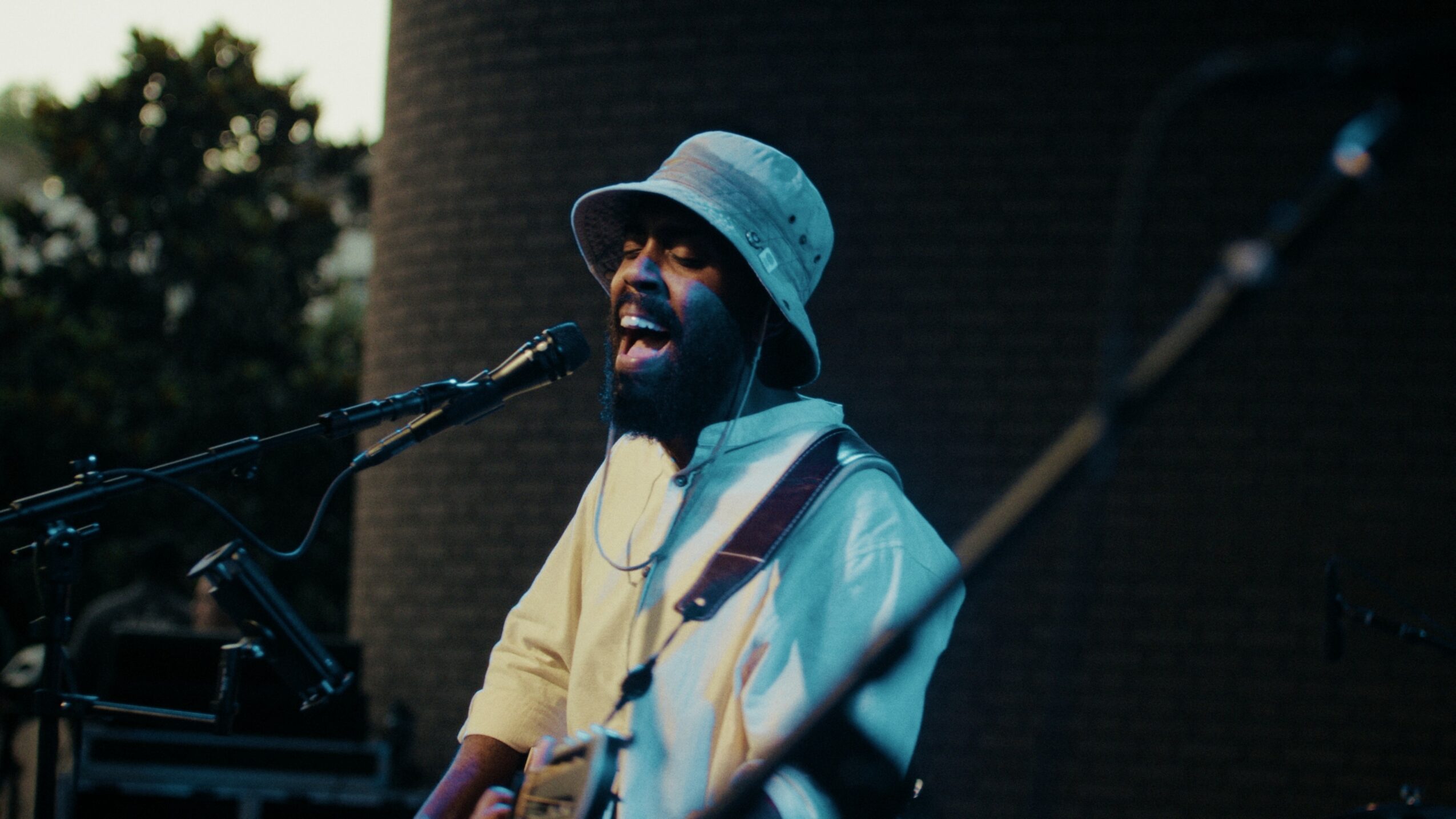Person singing into a microphone outdoors, wearing a light-colored hat and jacket, with trees in the background.