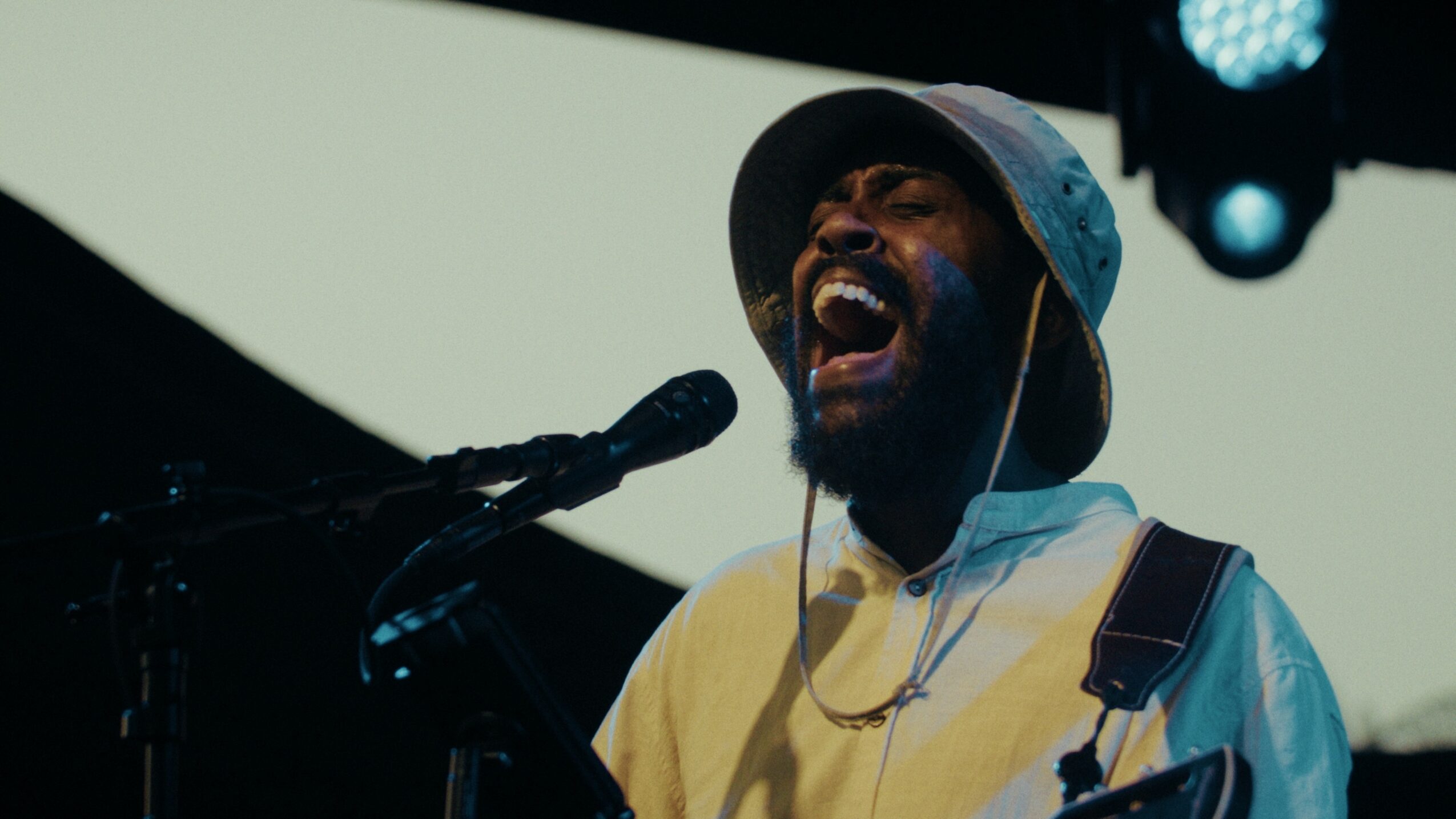Person singing passionately into a microphone with a keyboard nearby, wearing a hat and headphones, on stage.