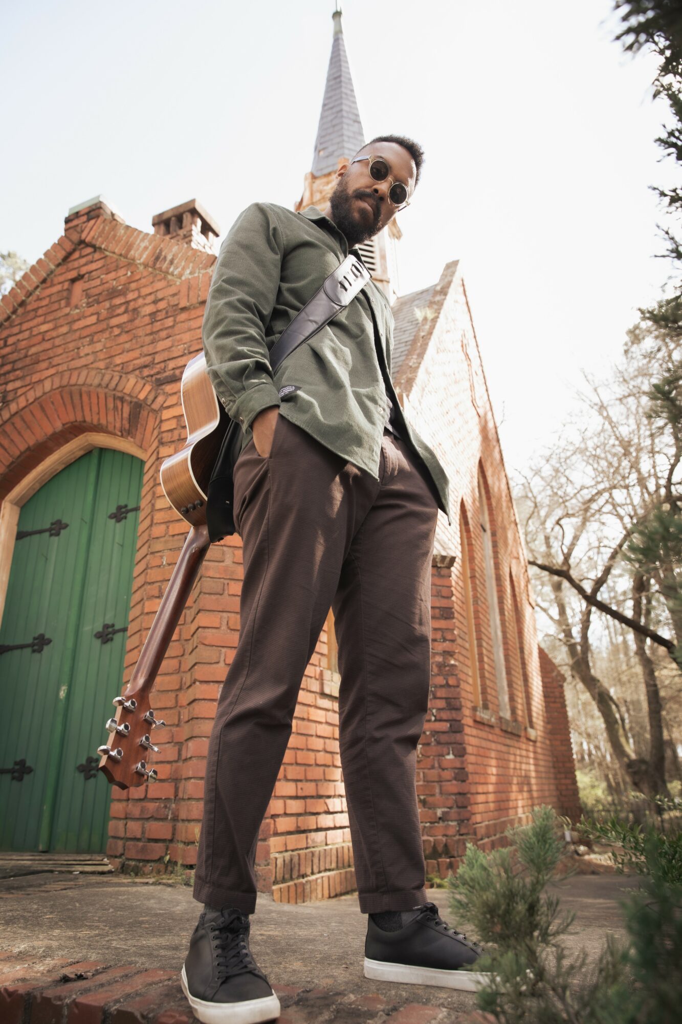 Man standing outside brick church with a guitar on his back, looking down, wearing sunglasses, green jacket, black pants, and sneakers.