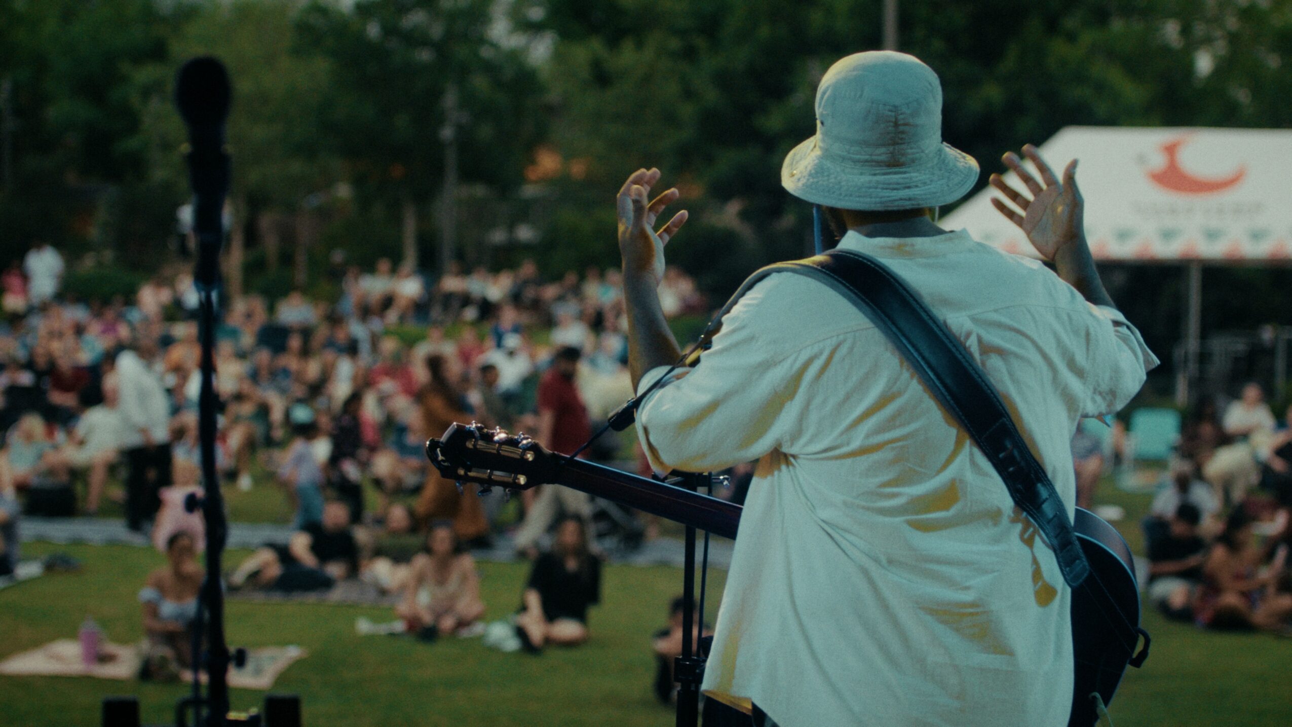 Person wearing a hat and white shirt playing guitar in front of a large crowd outdoors.