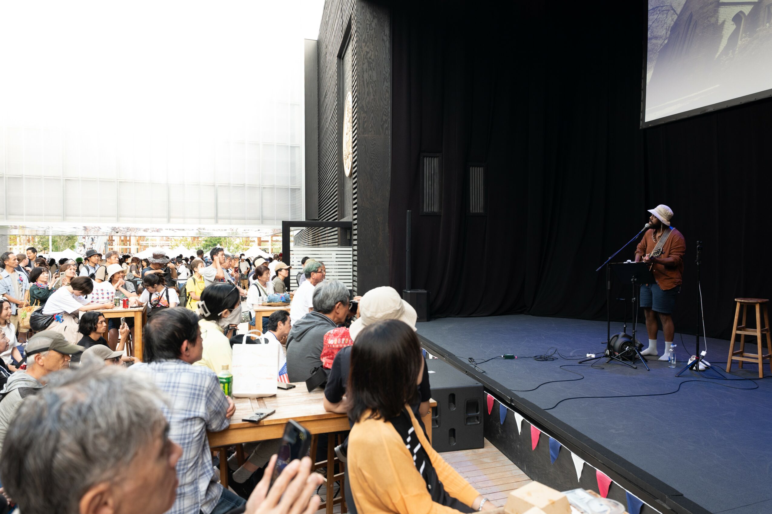 Person performing on stage with microphone in front of seated audience indoors, black curtain backdrop, some audience members visible.