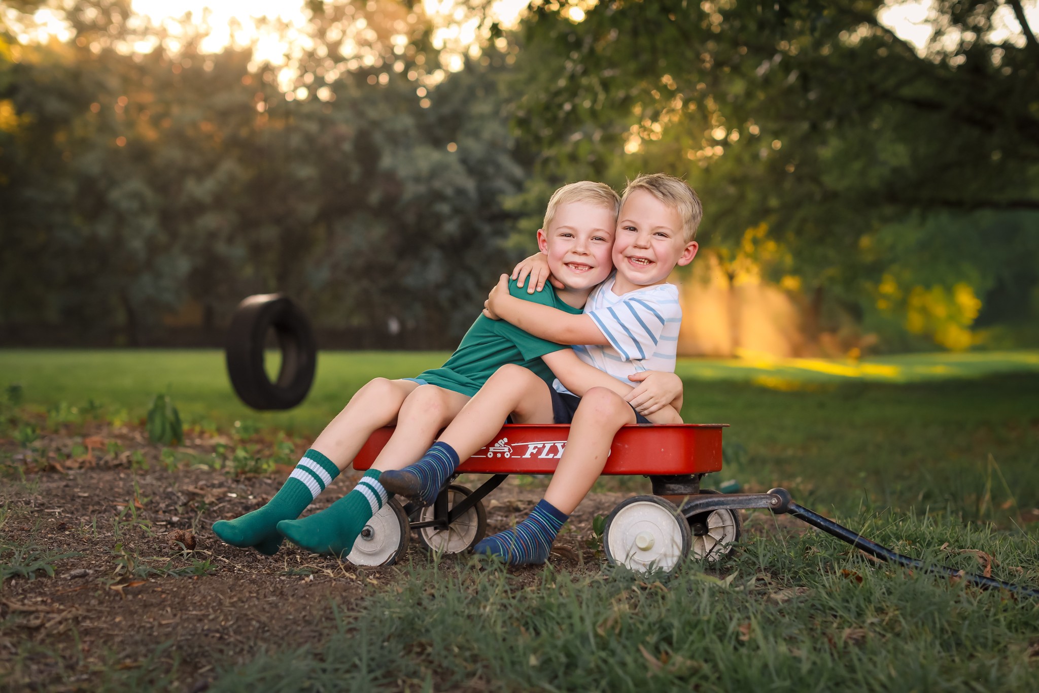 Two children sit on a red wagon outdoors, hugging and smiling, with trees and sunlight in the background.
