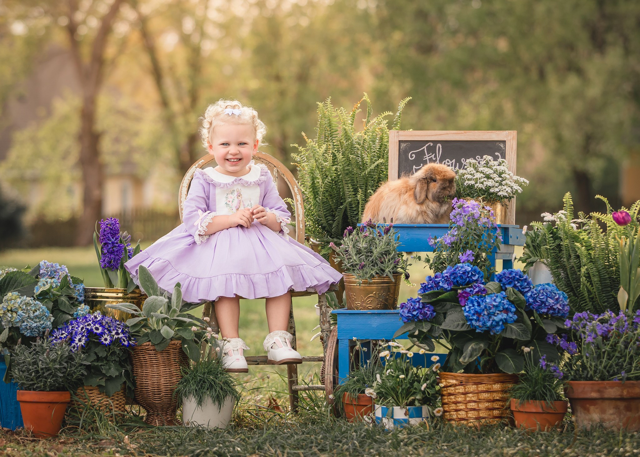 Young girl in a purple dress sitting among potted flowers outdoors with a puppy nearby, sunny day.