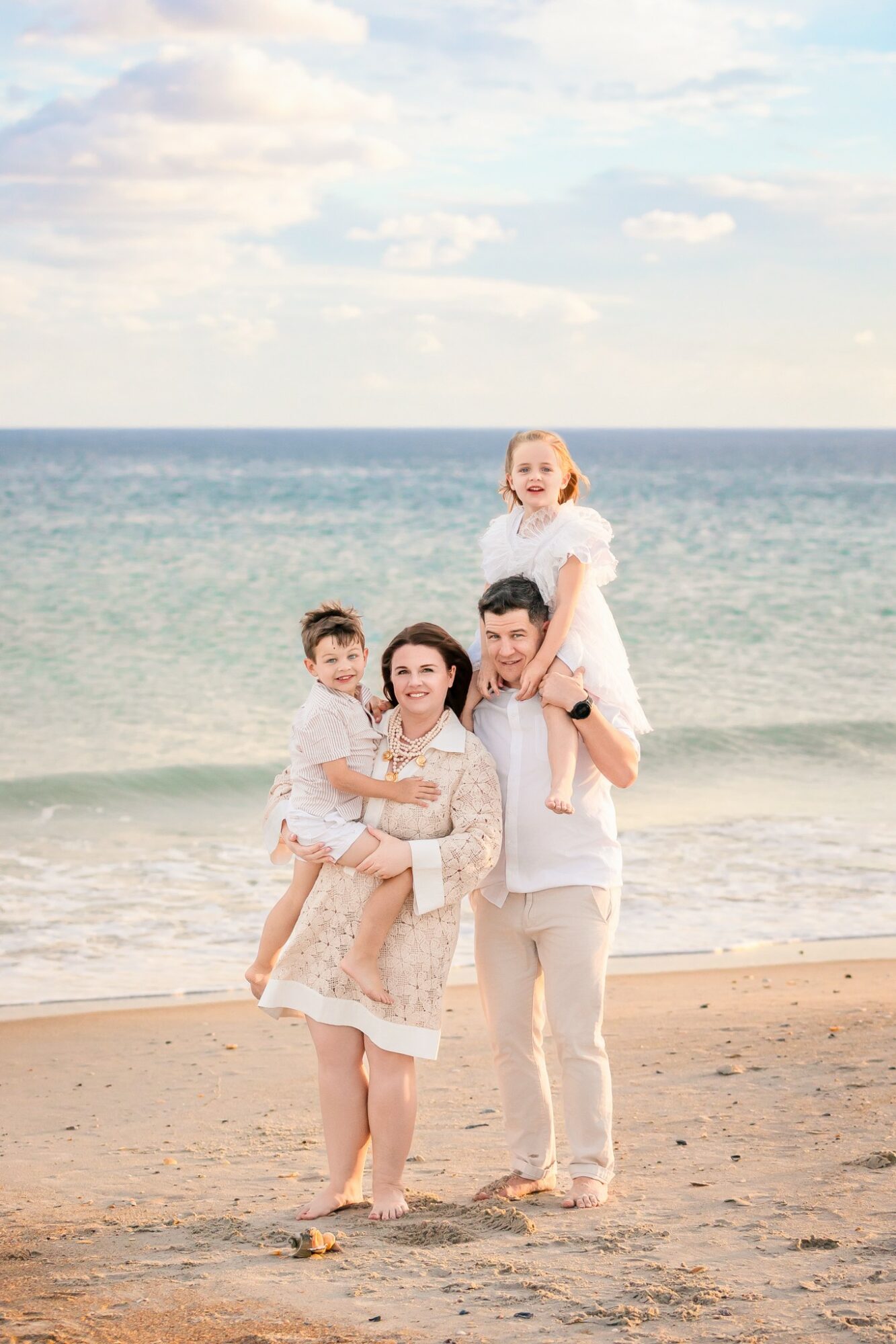 Family of four on beach, woman holding child, man carrying girl on shoulders, ocean and sky in background.