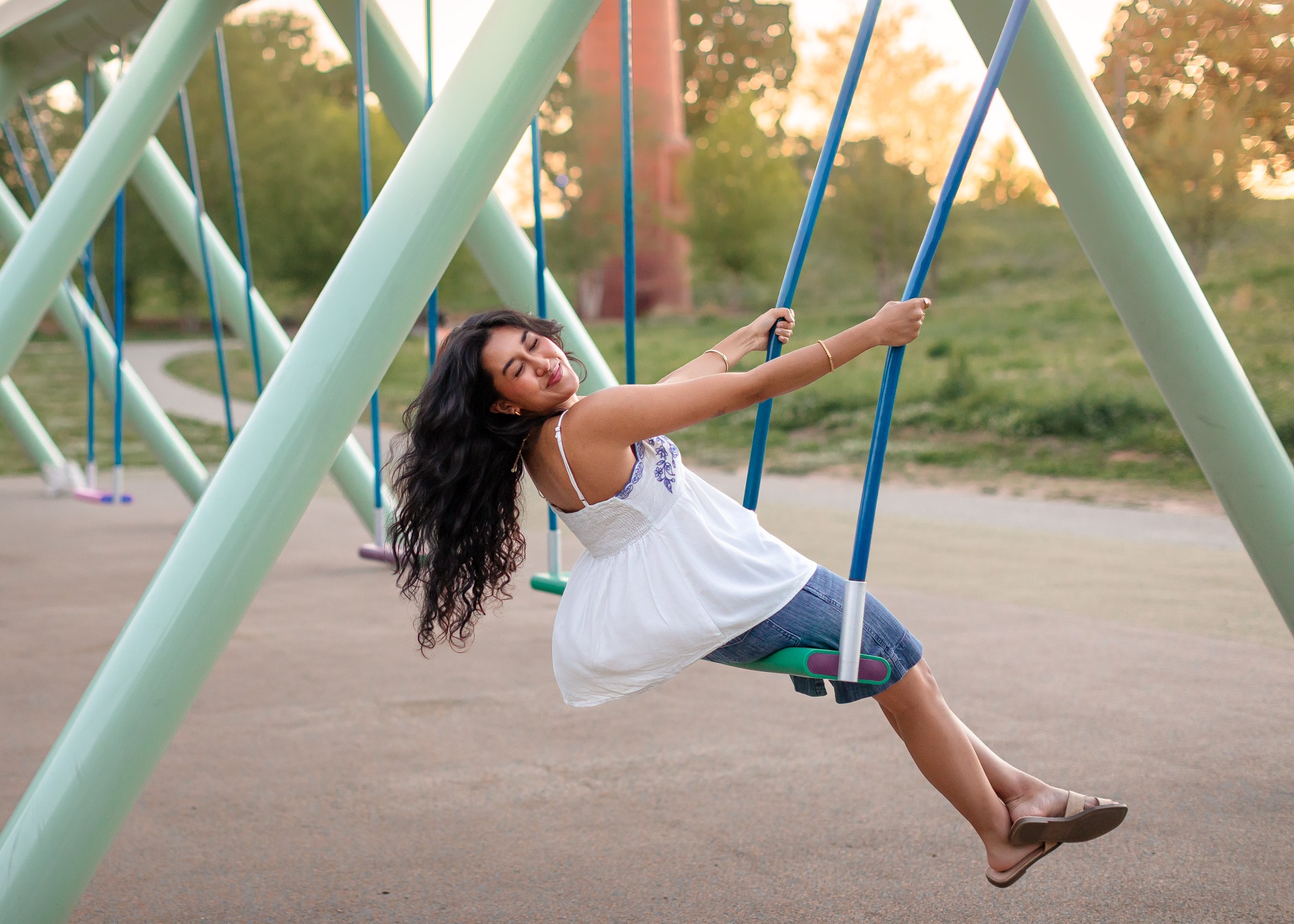 Girl swinging on a playground swing set outdoors during sunset, smiling and holding the swing's chains, wearing a white dress and jeans.