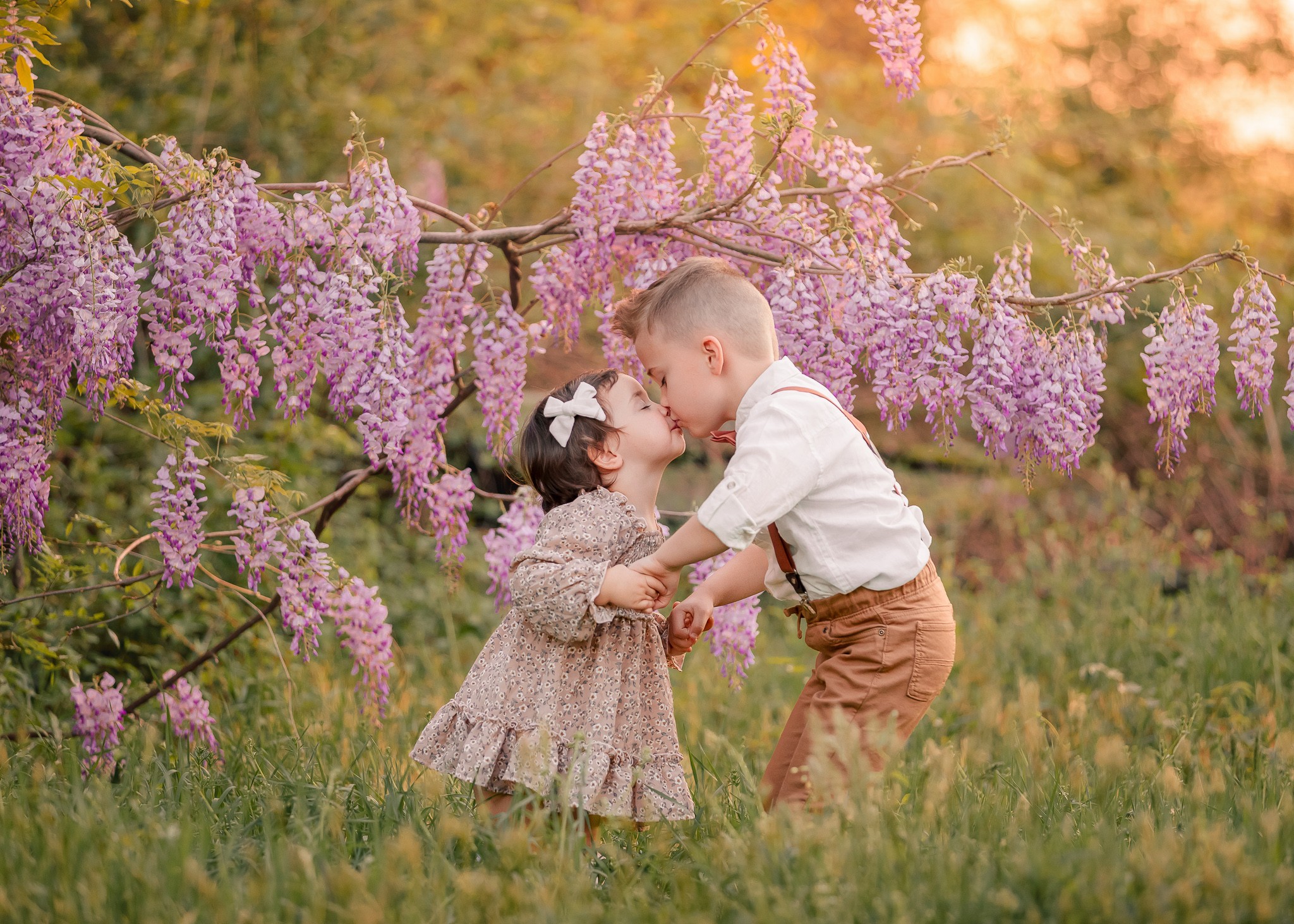 Two children, a boy and a girl, holding hands and playing under a flowering tree with pink blossoms outdoors.