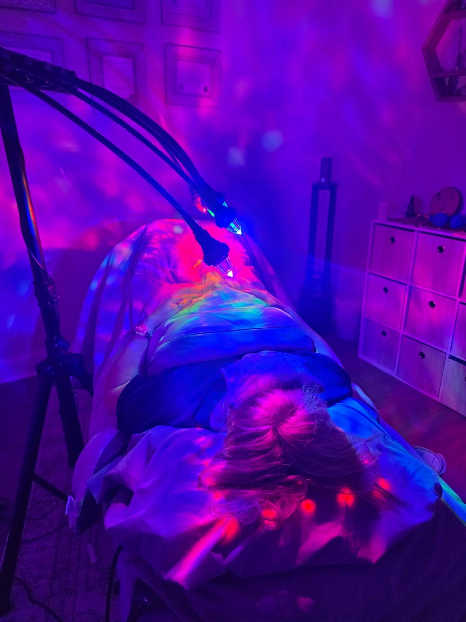 Person lying on a bed with colorful lights, in a room with furniture and a dresser, illuminated by vibrant lighting.