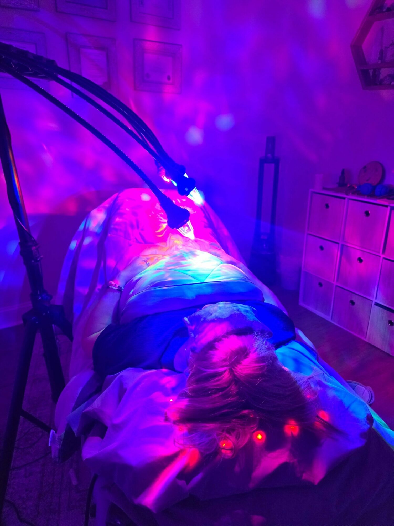 Person lying on bed with colorful lights, cables, and furniture in background, illuminated by purple and pink lighting.
