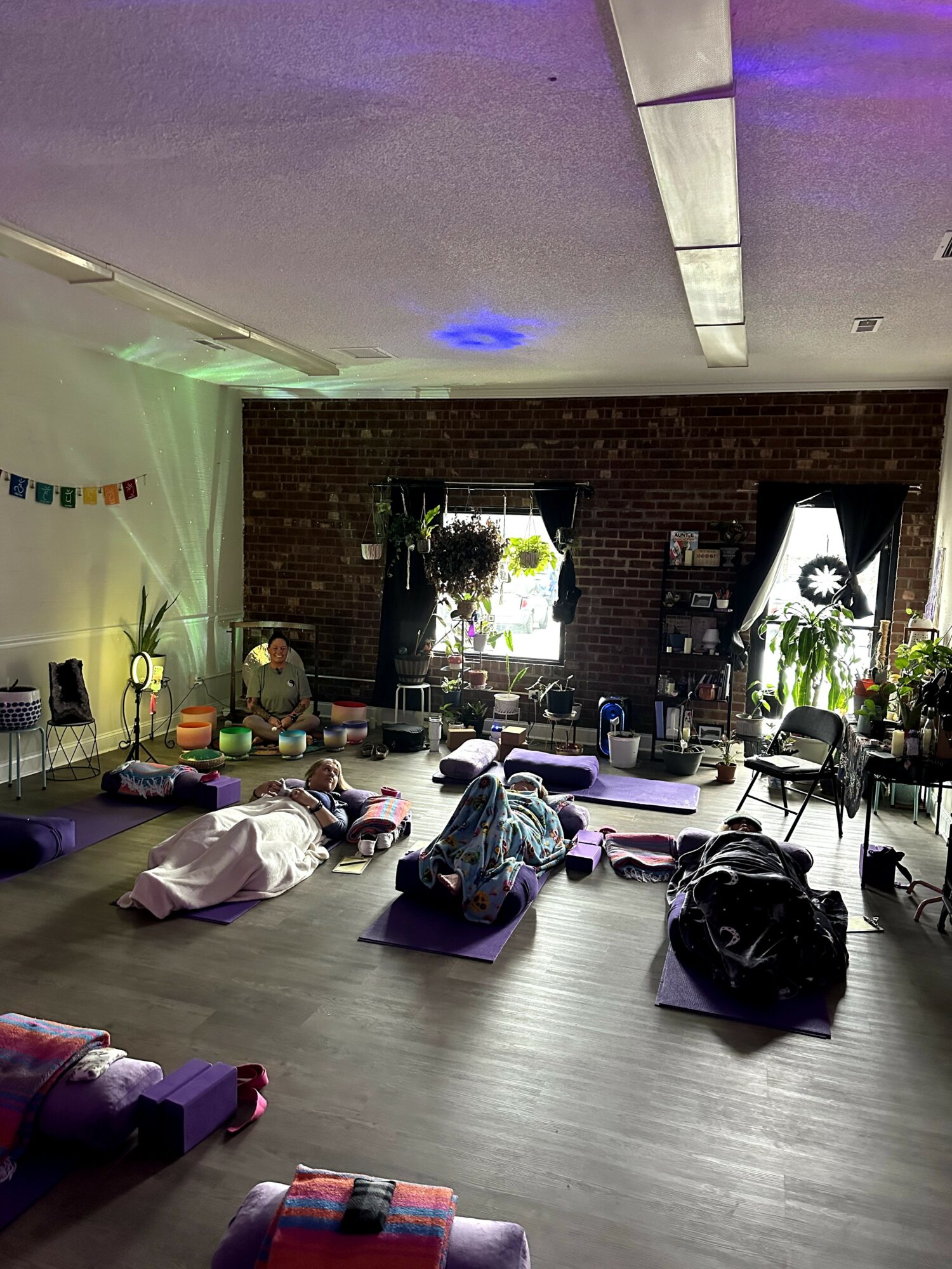 People practicing yoga on mats in a room with brick walls, windows, and plants, with soft lighting and decorations.