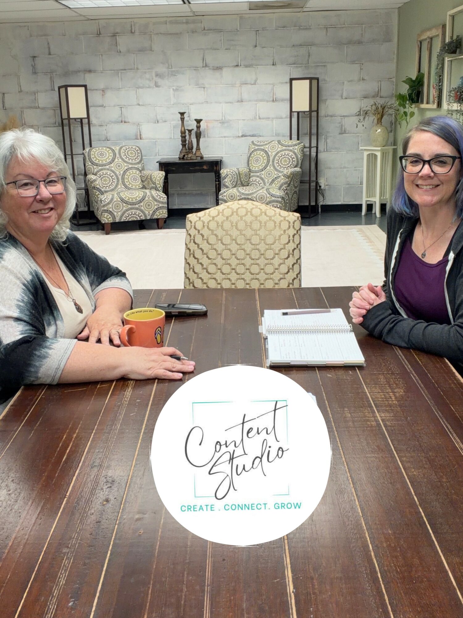 Two women sit at a wooden table in a room with a white brick wall, chairs, and lamps in the background.