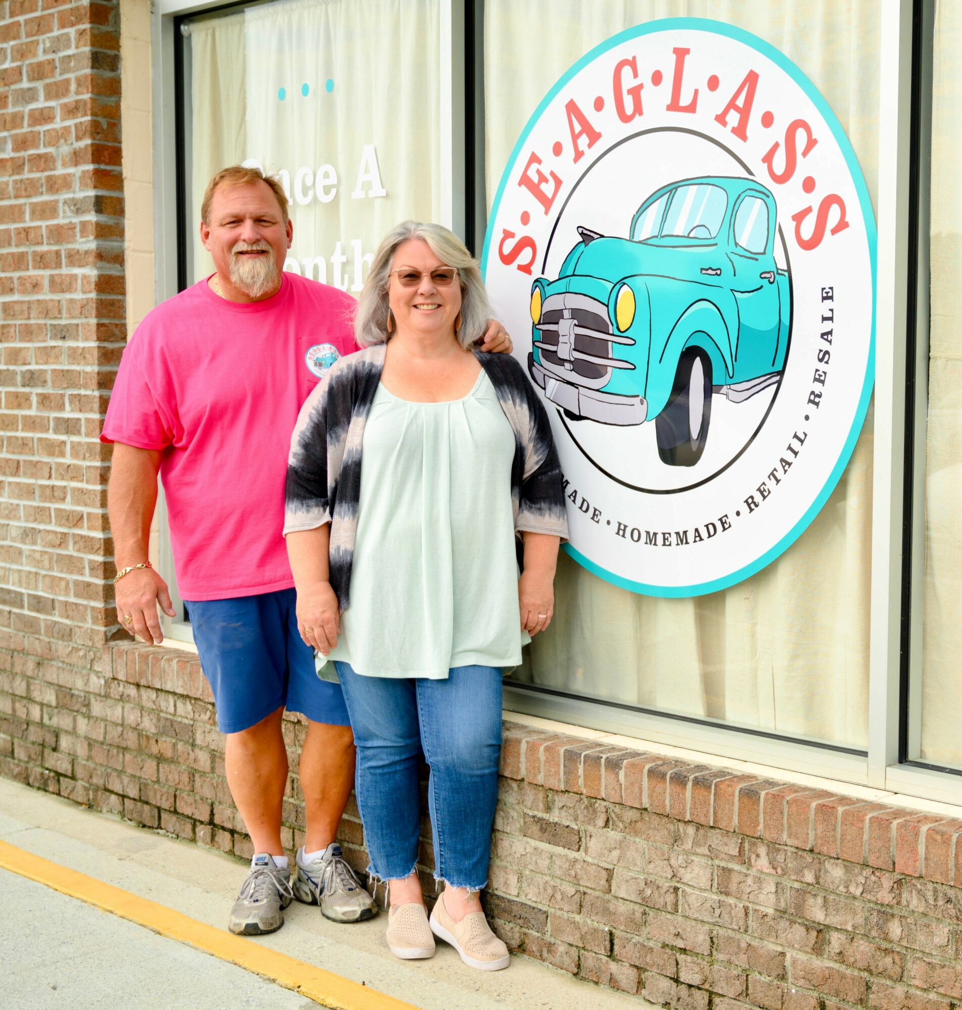 Two people standing outside in front of a window with a circular sign that reads 'S.E.A.G.L.A.S.S.' and features a blue vintage car.