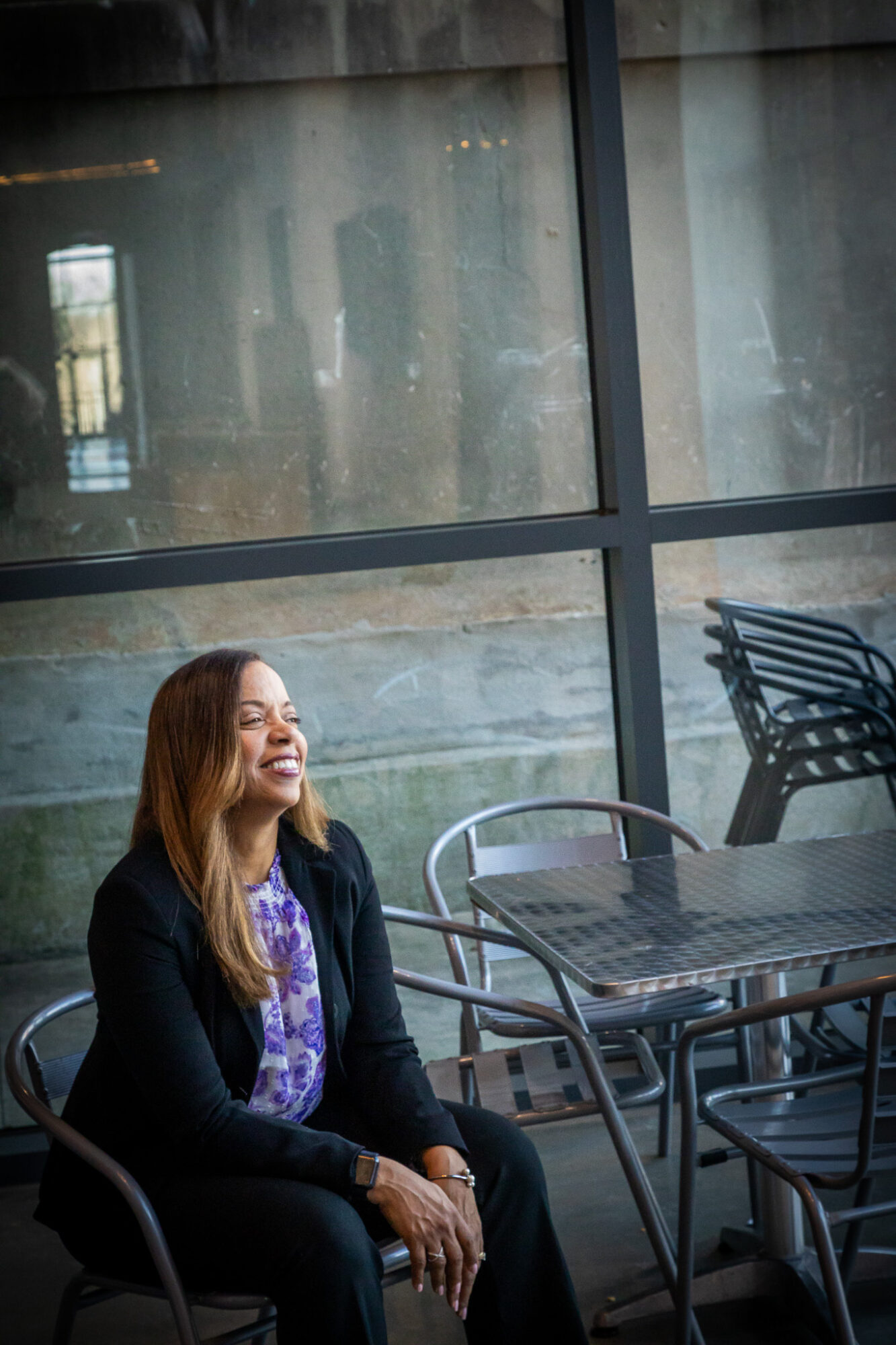 Woman with long hair sitting outdoors near large glass windows, smiling with closed eyes, surrounded by metal chairs and tables.