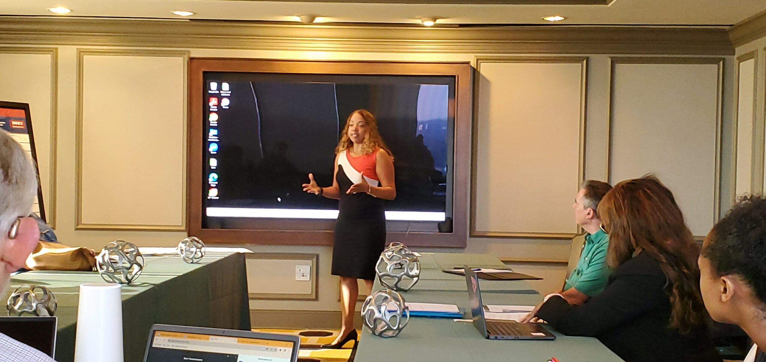 Woman standing in front of a large screen presenting to seated people in a conference room.