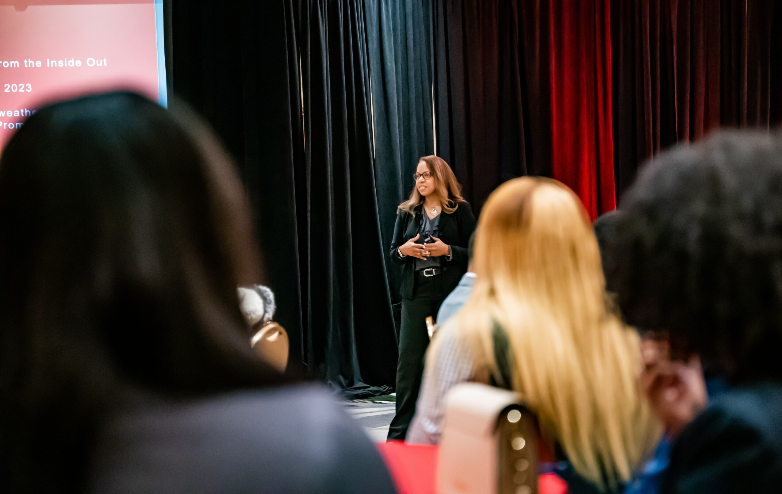 A woman stands on stage speaking to an audience in a room with black, blue, and red curtains.