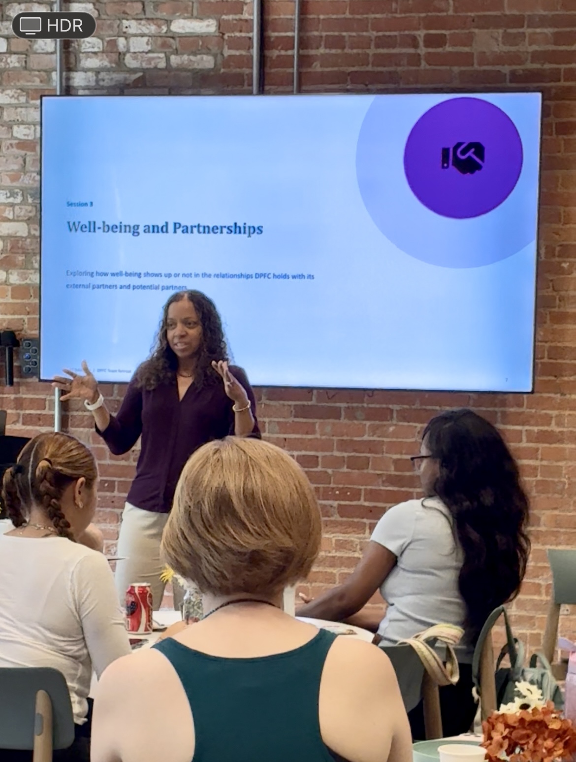 Woman presenting to a group of people in a room with a brick wall and a large screen displaying a presentation slide about well-being and partnerships.