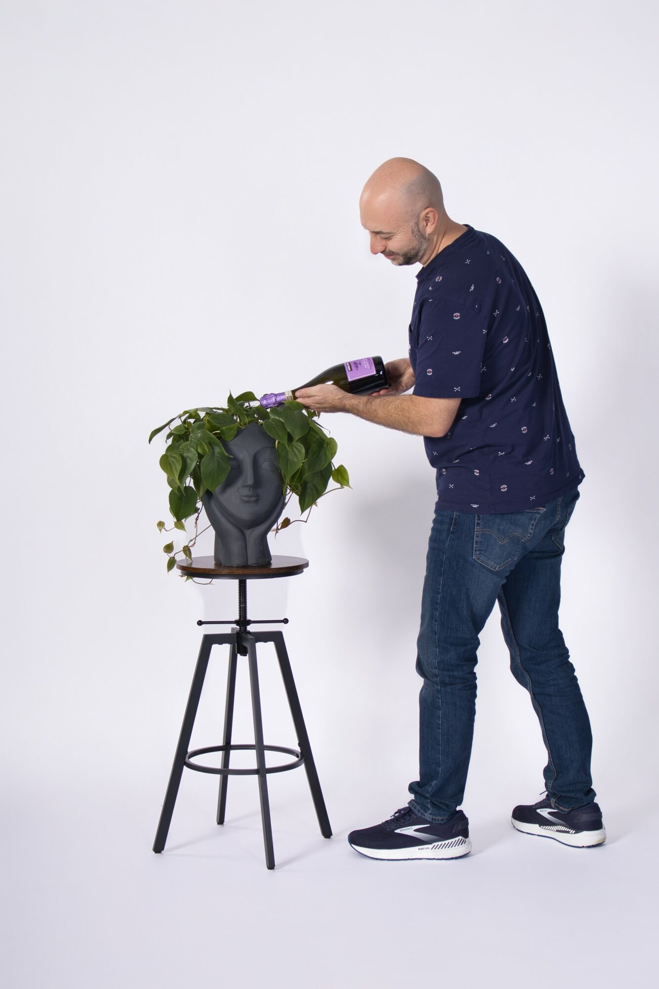 Man with a bald head, wearing a navy shirt and jeans, looks at a tablet while standing next to a potted plant on a stand.