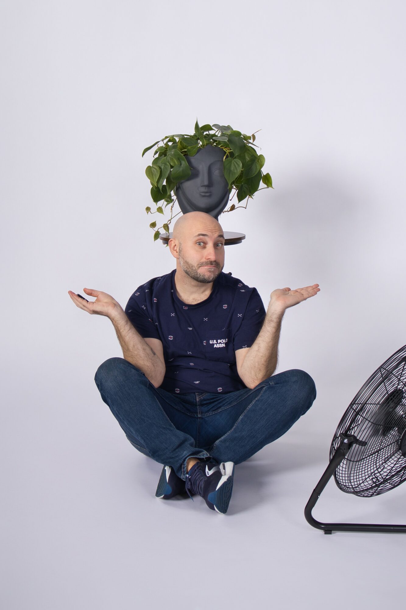 Man sitting cross-legged with arms raised, a plant on his head, and a fan nearby, against a plain white background.