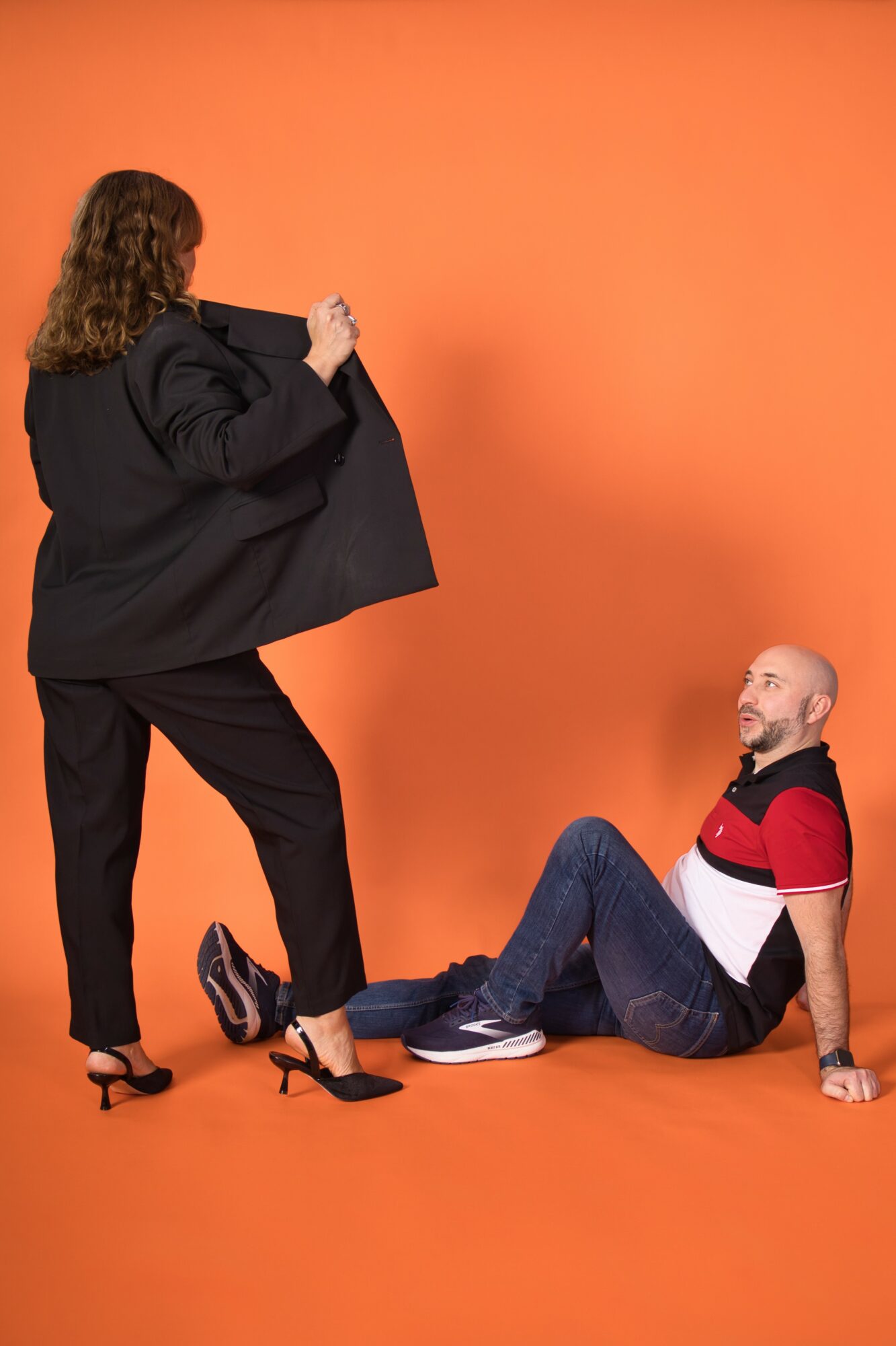 Woman standing with a large black bag, man sitting on the floor, both against an orange background.