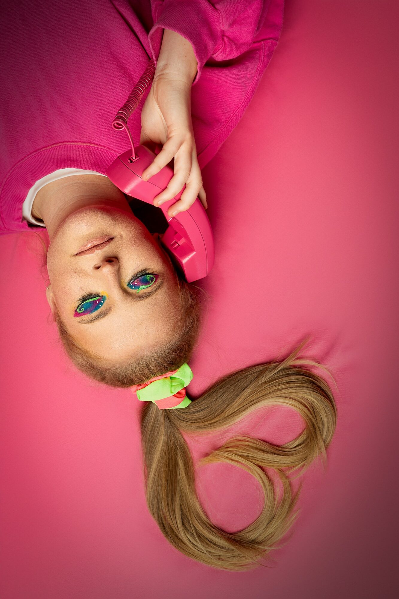 Young girl with a ponytail, wearing pink, lying on pink surface, talking on a pink phone, smiling.