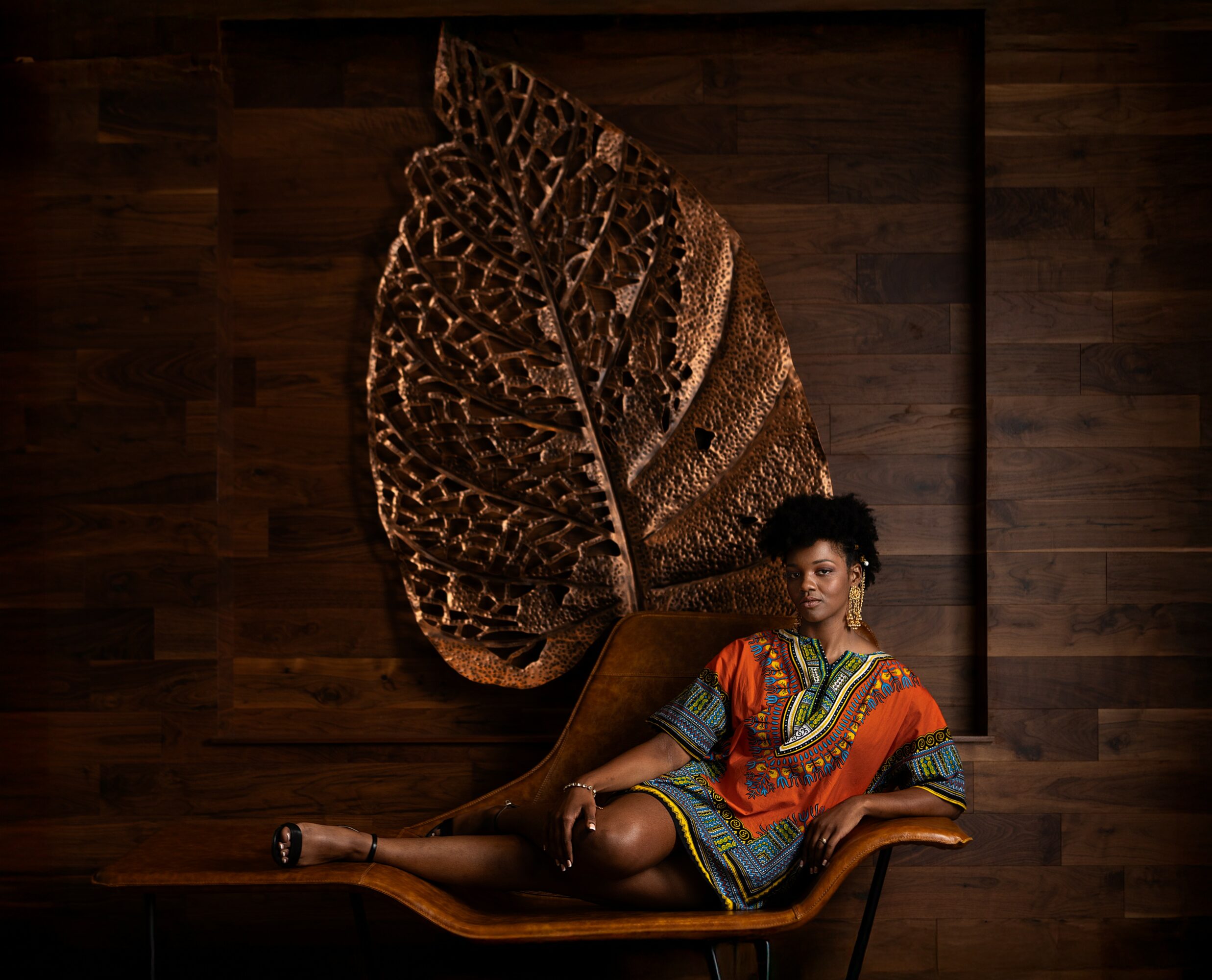 Woman in colorful dress reclining on a chair in front of a large leaf-shaped artwork on a wooden wall.