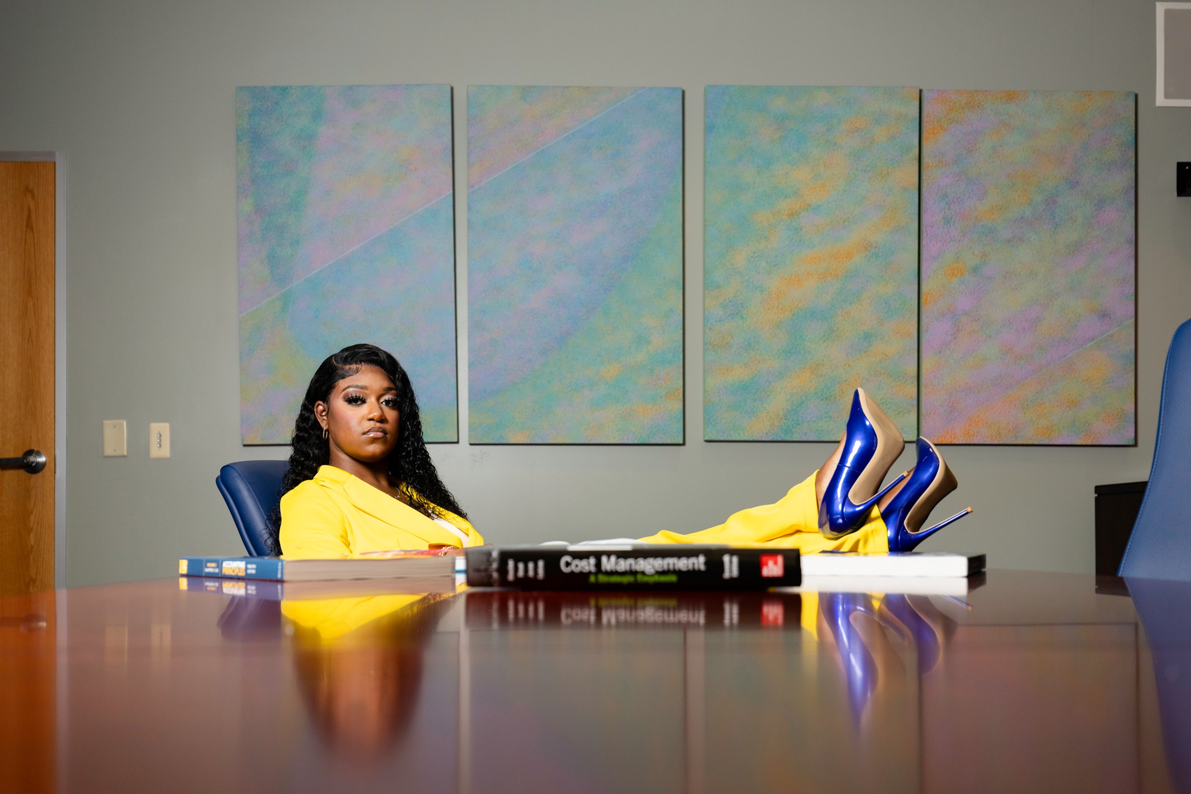 Woman in yellow outfit and blue shoes reclining at a conference table with abstract artwork behind her.