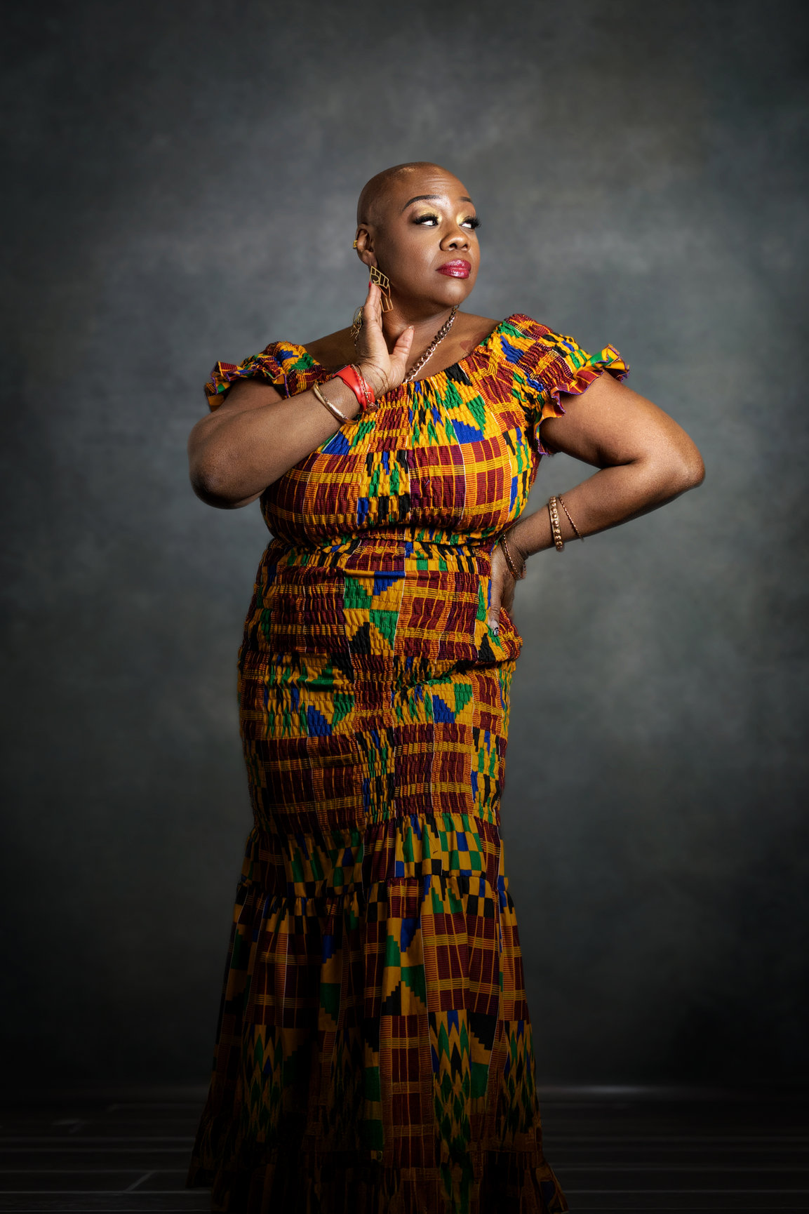 Woman in colorful traditional dress poses with hand near face against dark background.