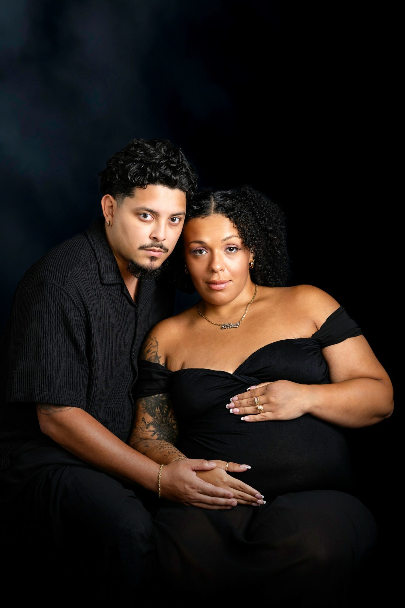 A man and a pregnant woman sit close together against a dark background, with the woman resting her hand on her belly.