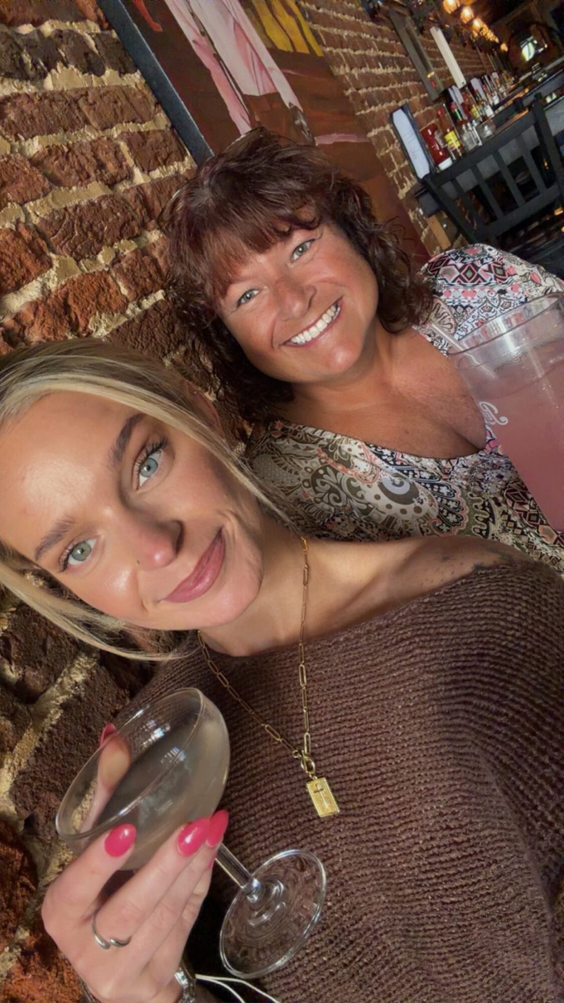 Two women smiling, one holding a glass, in a cozy indoor setting with brick wall background.