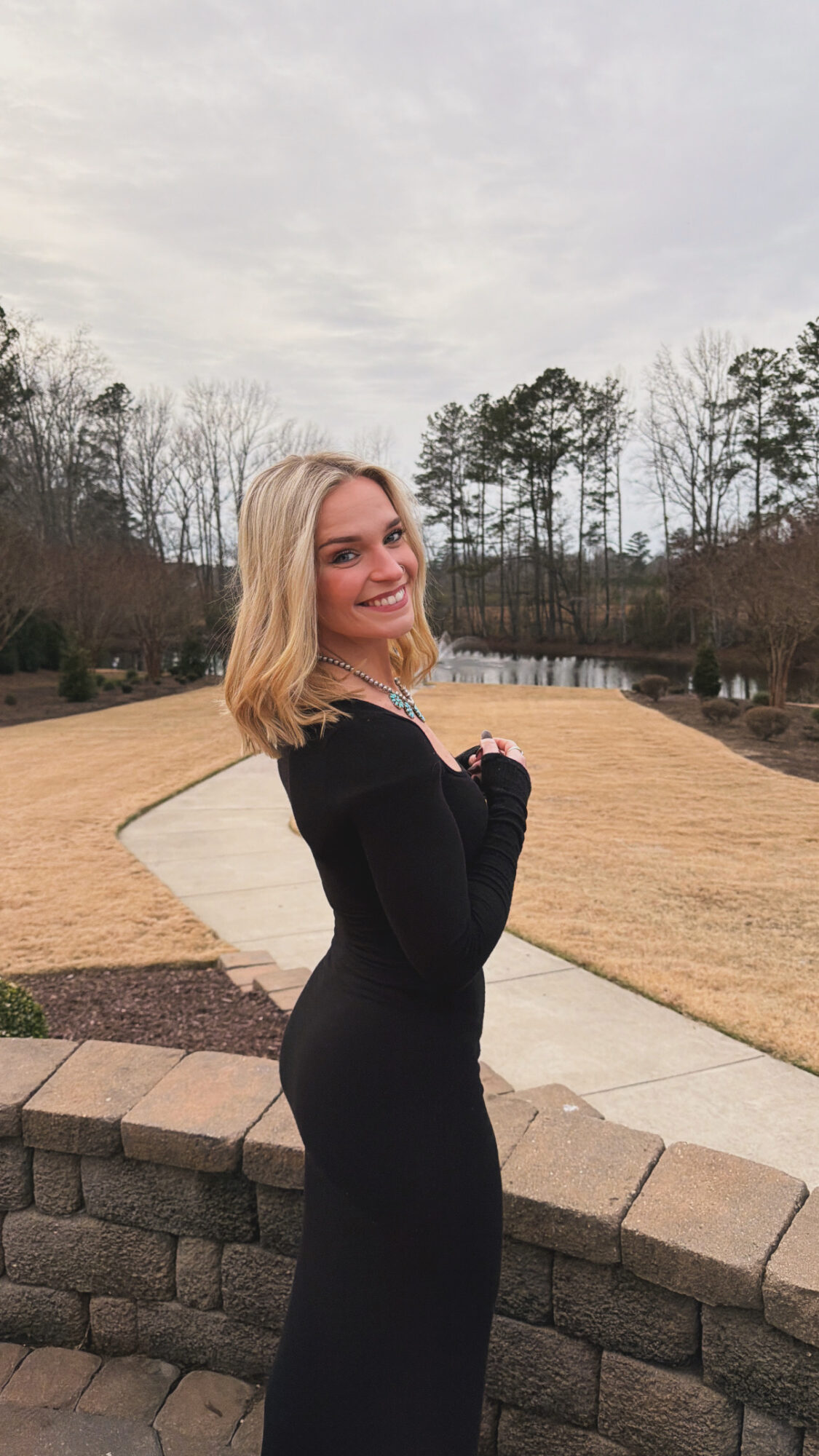 Young woman with blonde hair in a black dress standing outdoors on a curved stone wall, smiling, with trees and cloudy sky behind her.