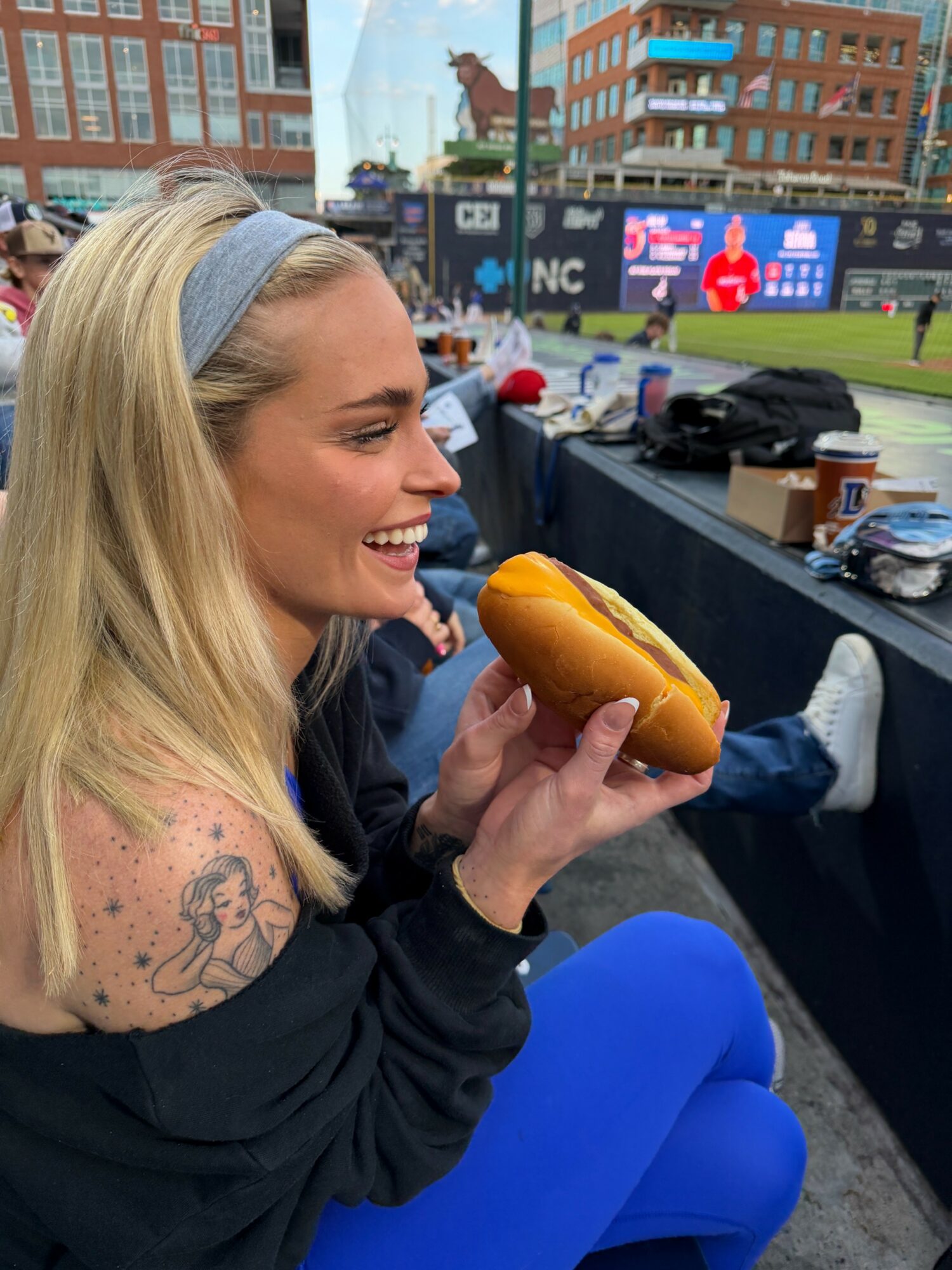 Young woman with blonde hair and a tattoo on her arm smiling and holding a hot dog at an outdoor event.