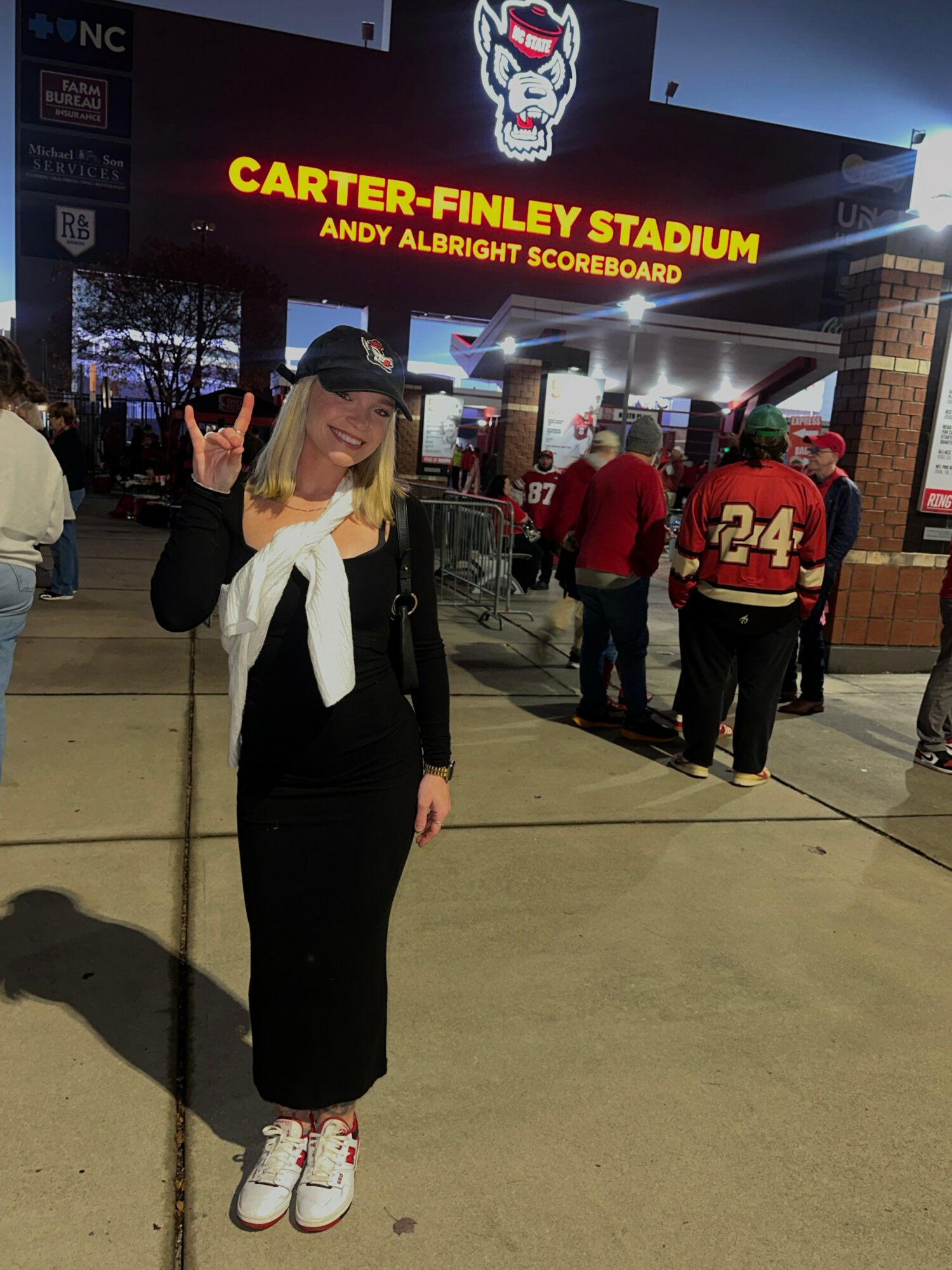 Woman in black outfit and cap making a hand gesture outside Carter-Finley Stadium at night.