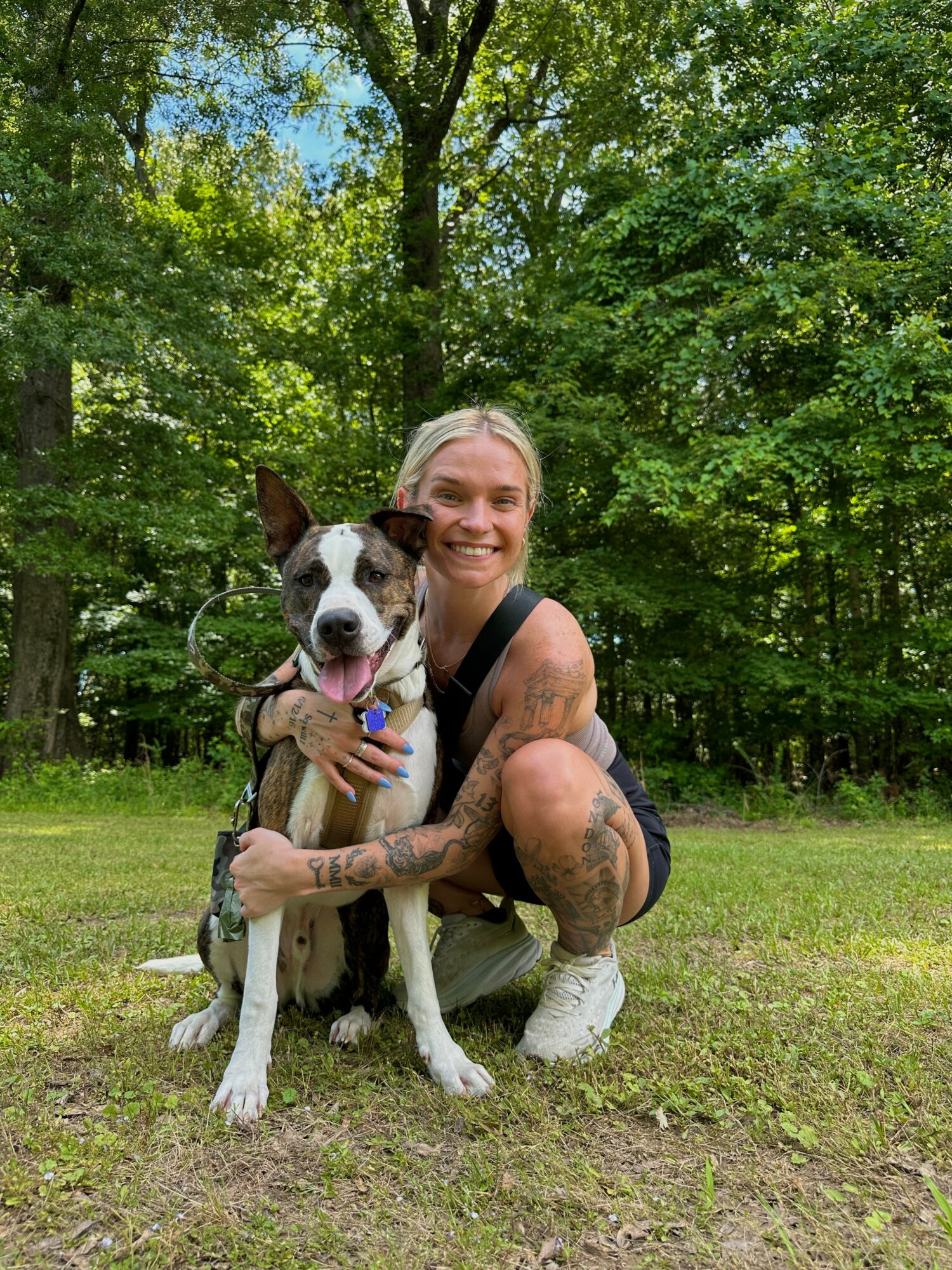 Woman crouching outdoors with a black and white dog in a grassy area surrounded by trees, smiling at the camera.