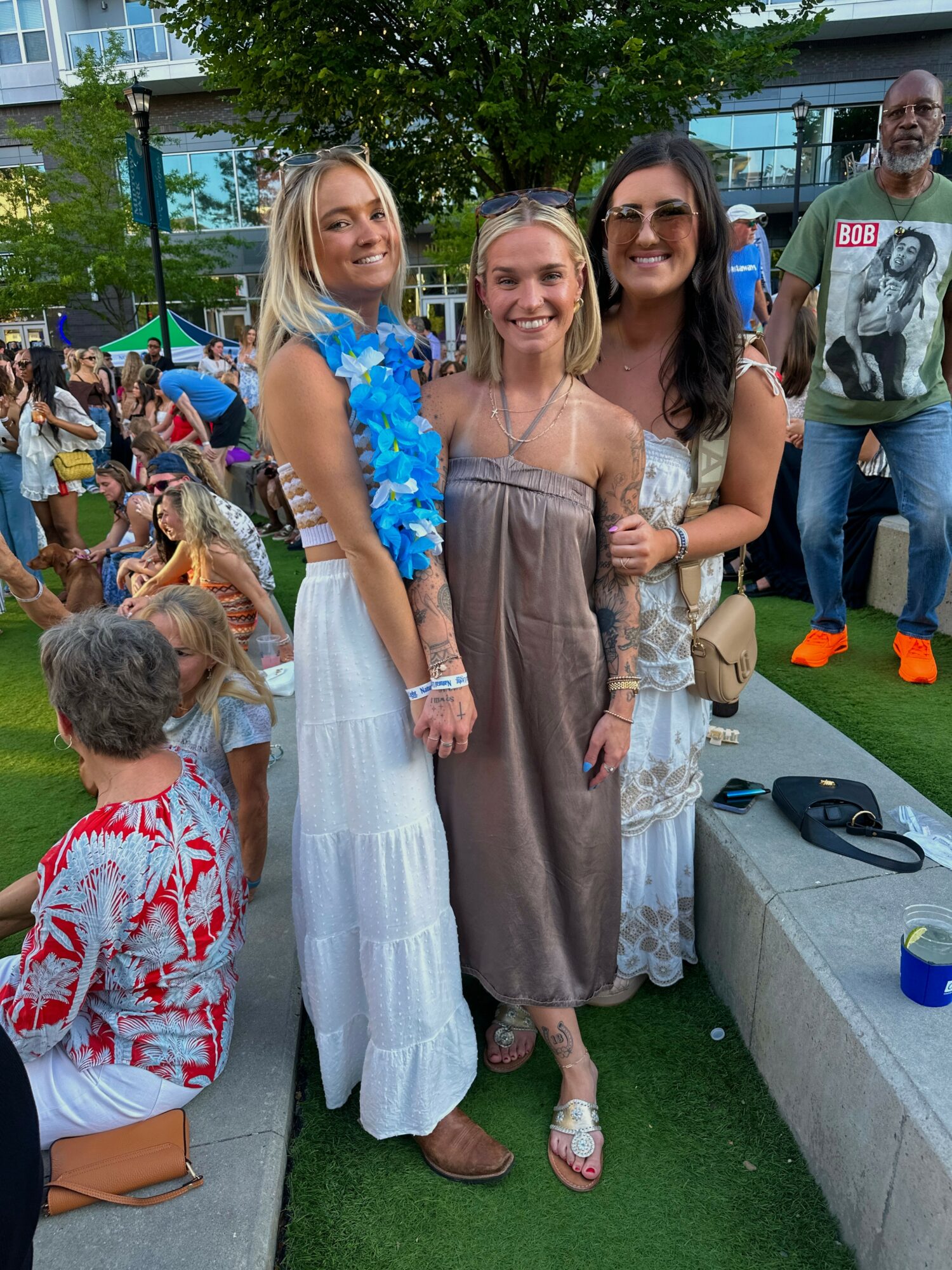 Three women standing outdoors, smiling, with a crowd and trees in the background.