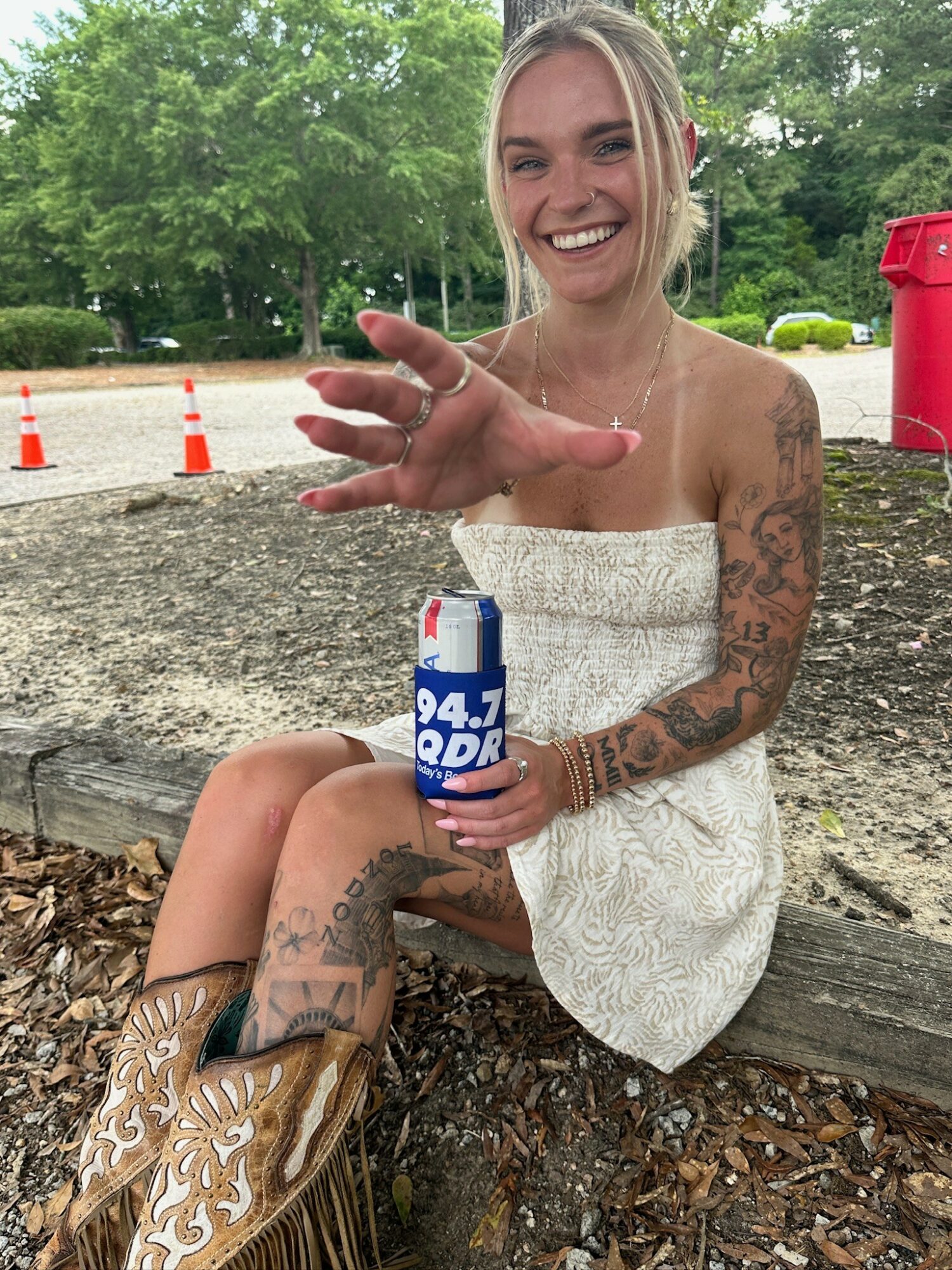 Smiling woman with tattoos sitting outdoors, holding a soda can, reaching out with her hand, wearing a white dress and cowboy boots.