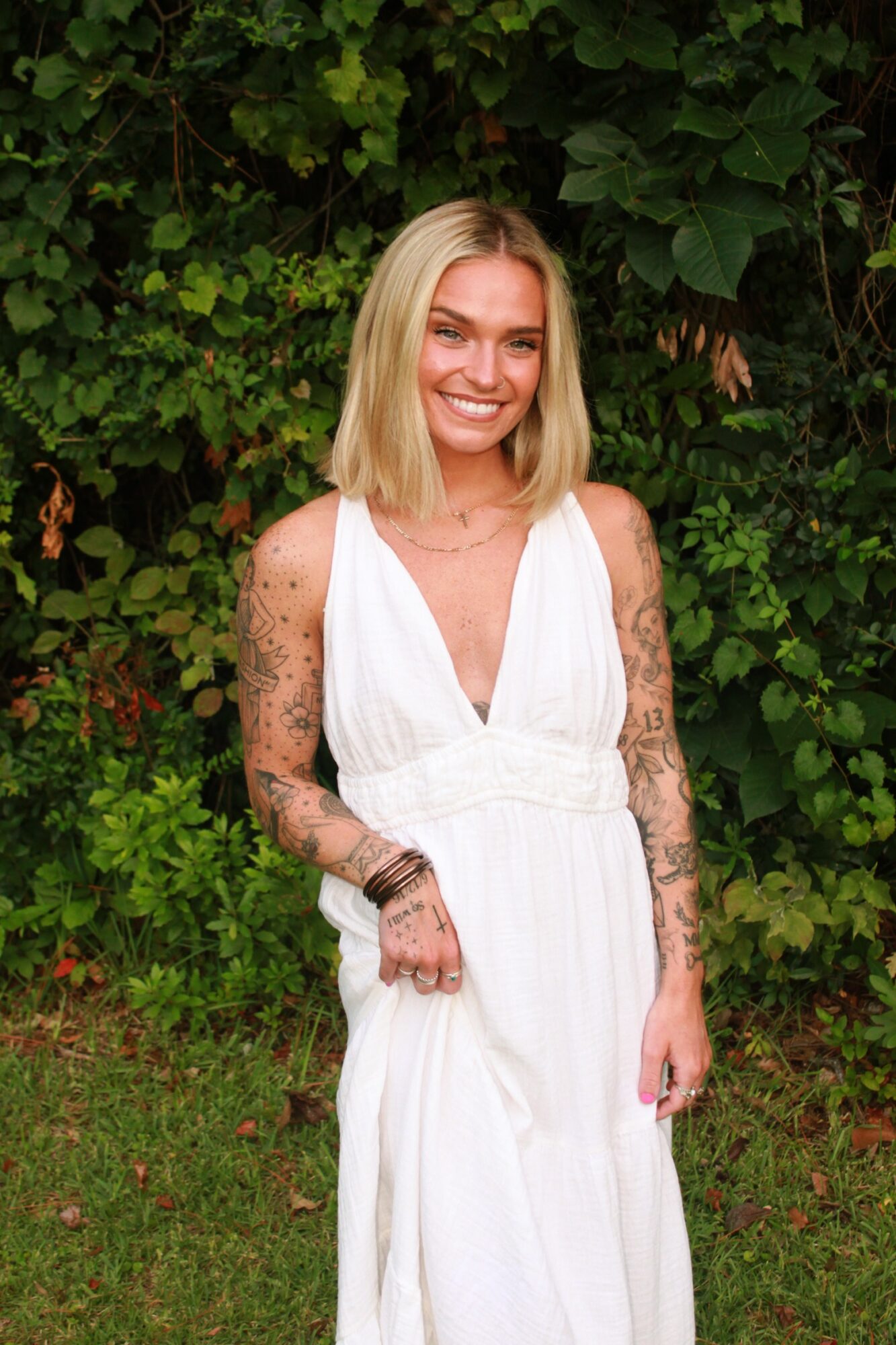 Young woman with blonde hair in a white dress standing outdoors with green foliage background.